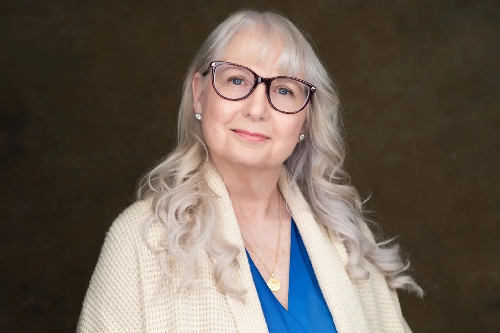 A smiling woman with long blonde hair, wearing a purple patterned blouse and a gold necklace, against a plain light blue background.