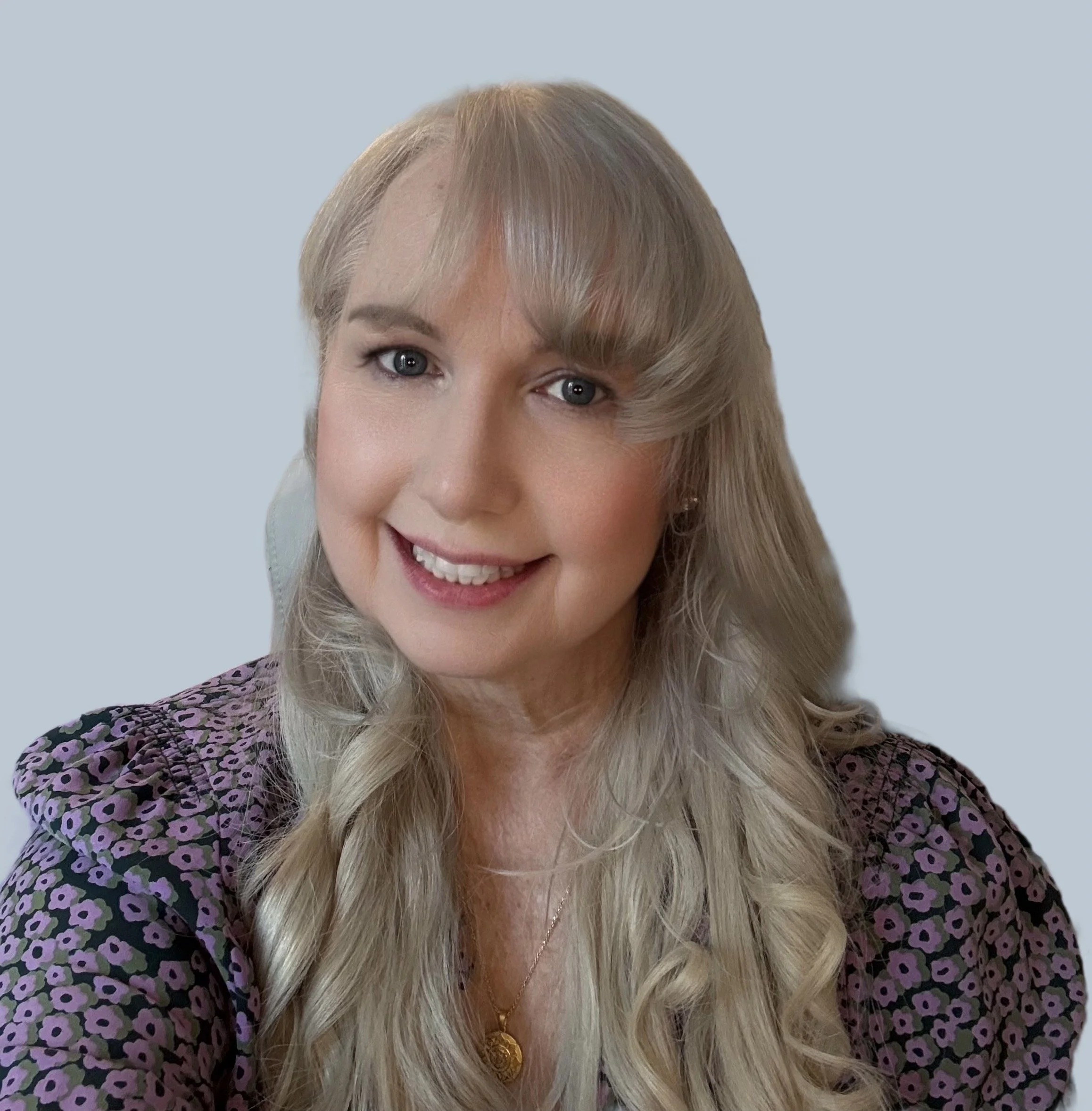 A smiling woman with long blonde hair, wearing a purple patterned blouse and a gold necklace, against a plain light blue background.