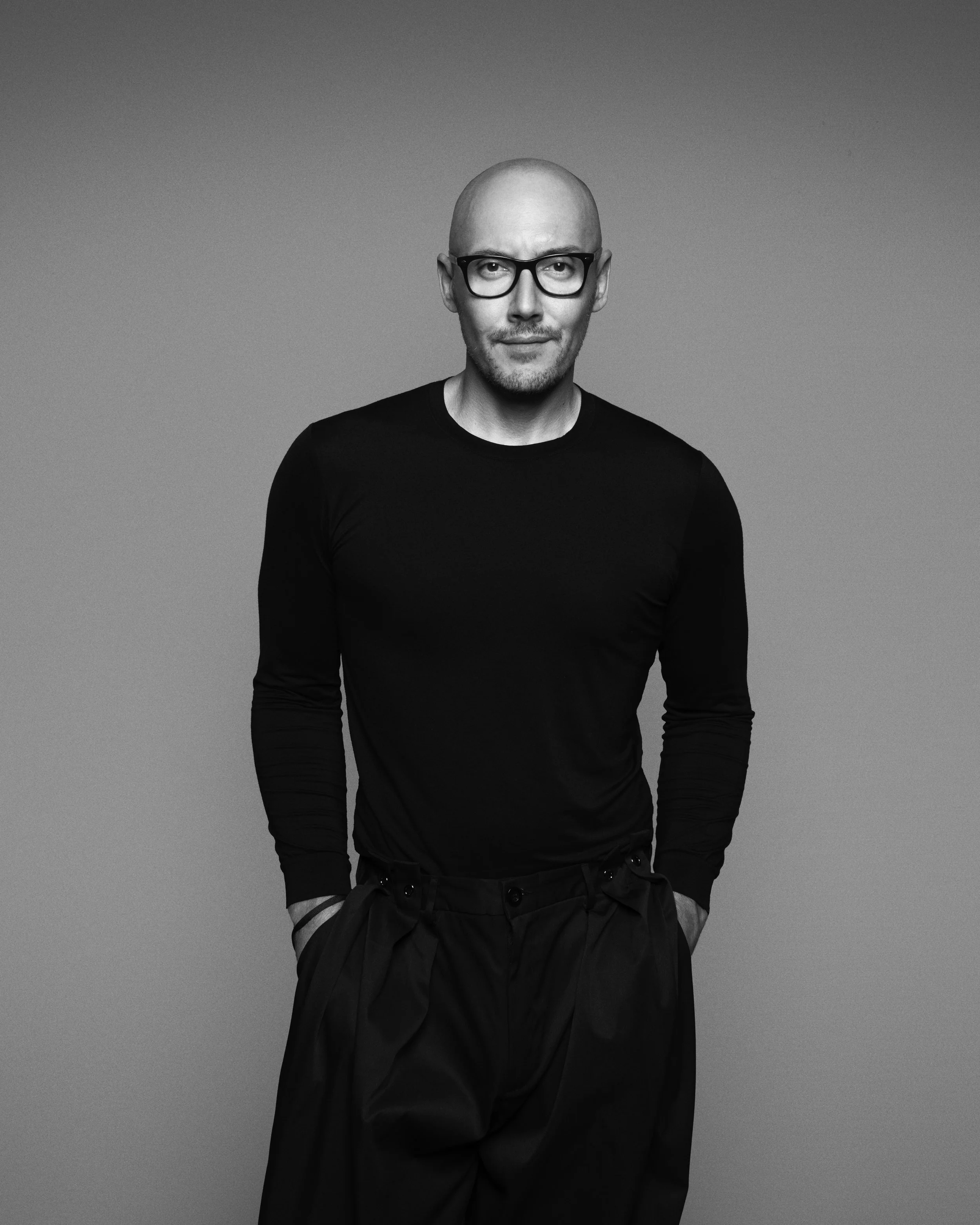 Black and white portrait of a man wearing glasses, a long-sleeved black shirt, and black pants, standing with his hands in his pockets against a plain background.
