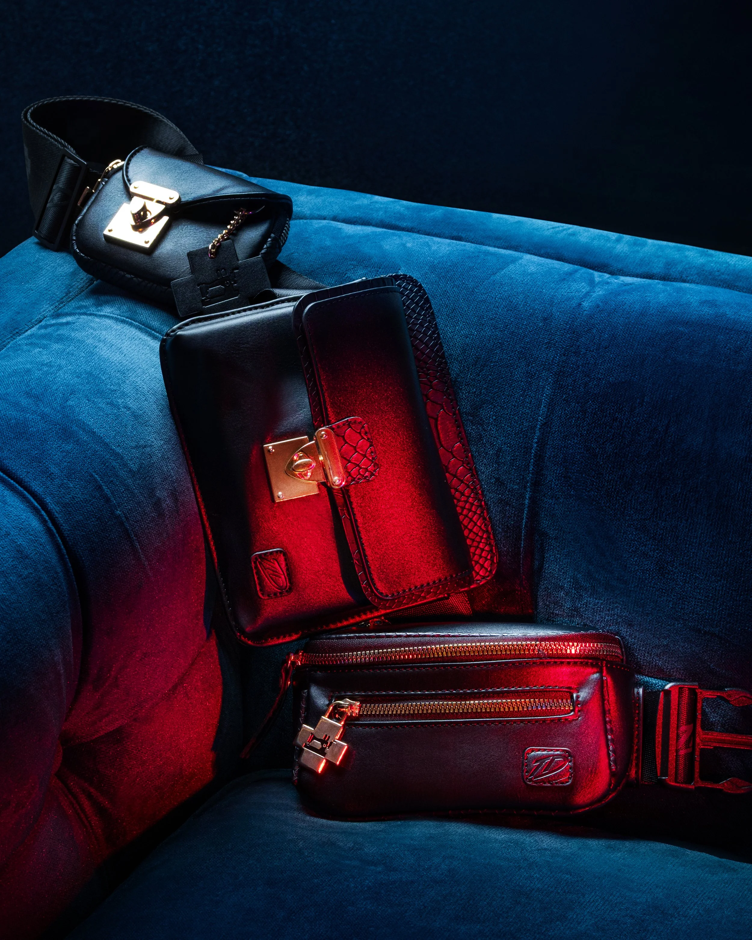 Close-up of a black handbag with gold hardware, red accents, and a zipper, resting on a blue fabric surface, with the background dark and plain.