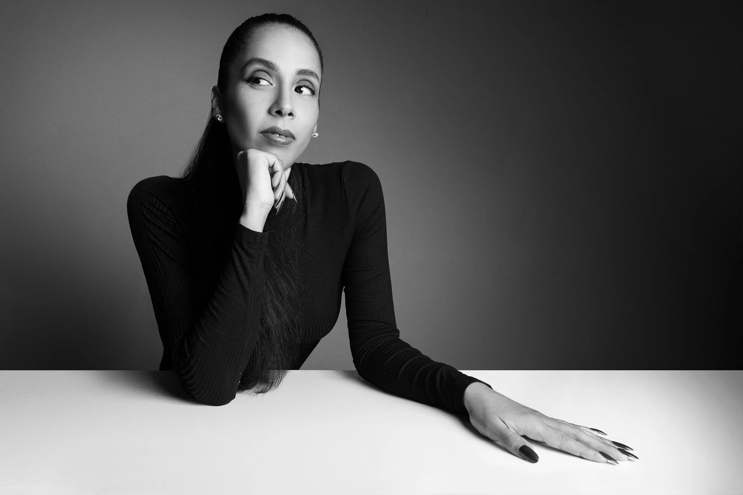 Black and white photograph of a woman with long dark hair, posed with her hand under her chin and her left arm extended on a white surface, wearing a tight long-sleeve black top, against a dark background.