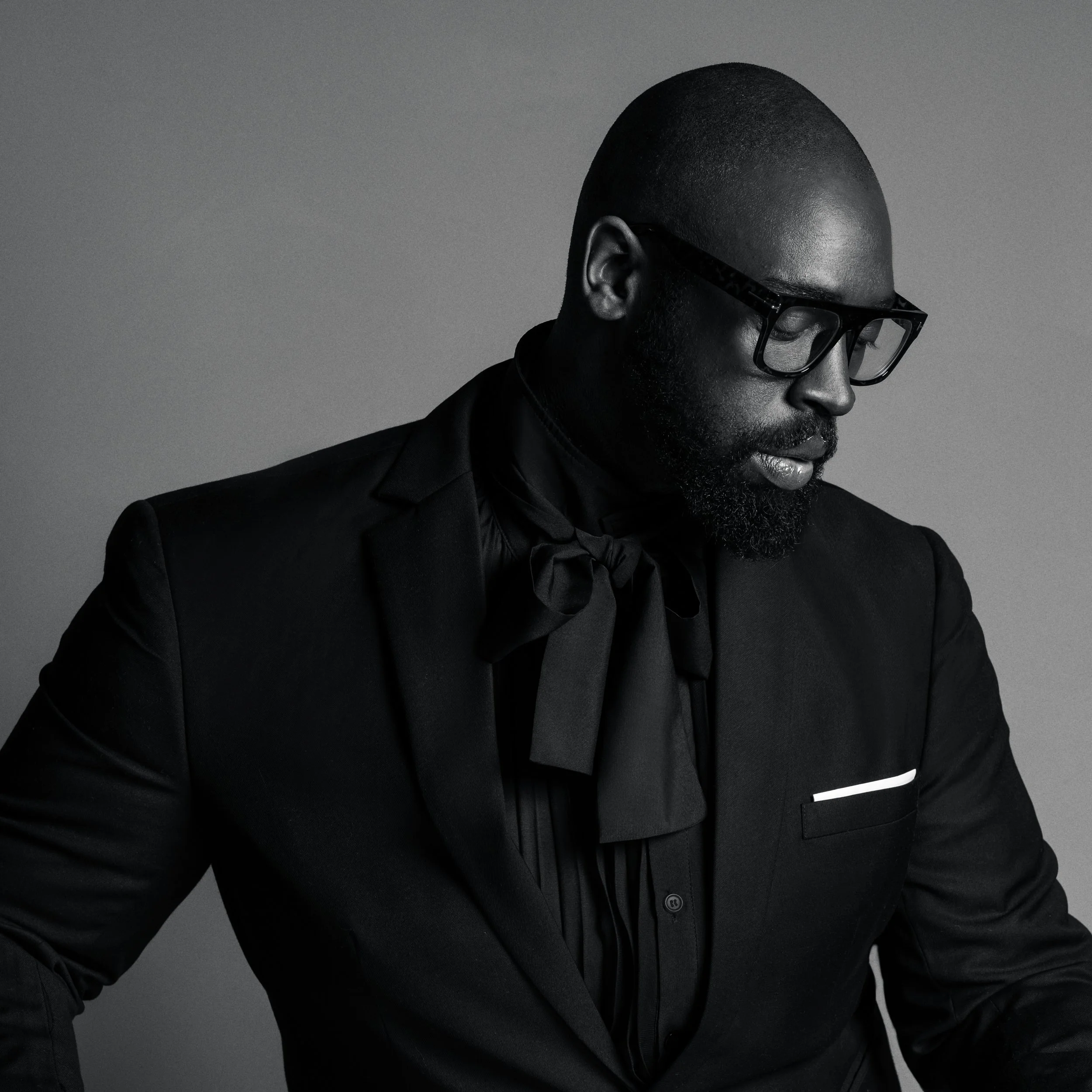A man with a shaved head wearing black-framed glasses, a black suit, and a black shirt, looking down against a gray background.