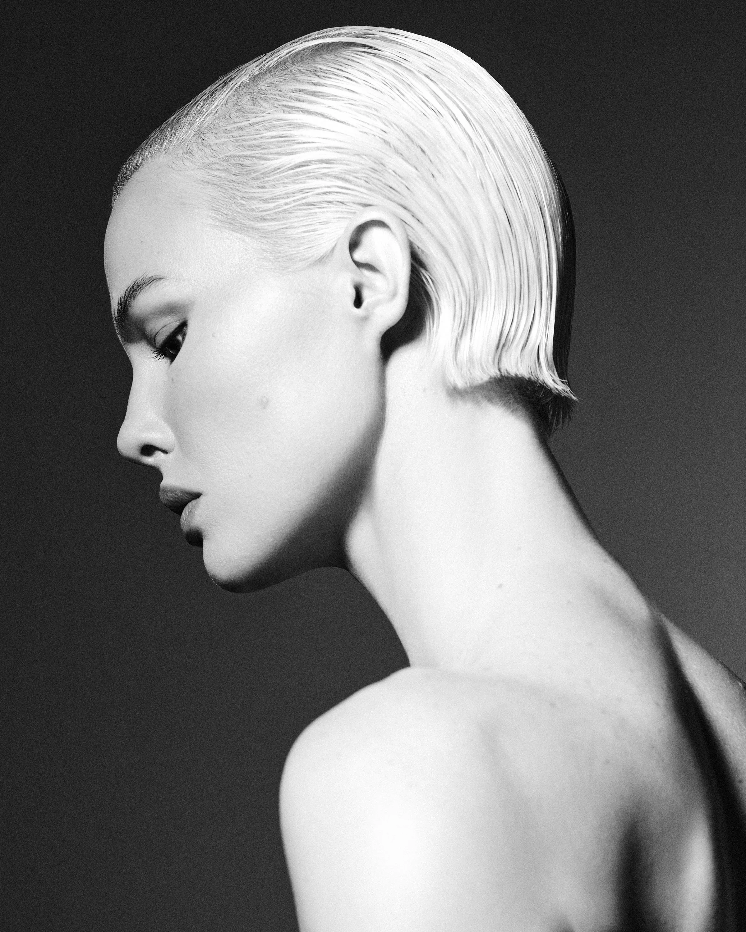 Black and white close-up profile of a woman with platinum blonde hair slicked back, showing her face, ear, and neck against a dark background.