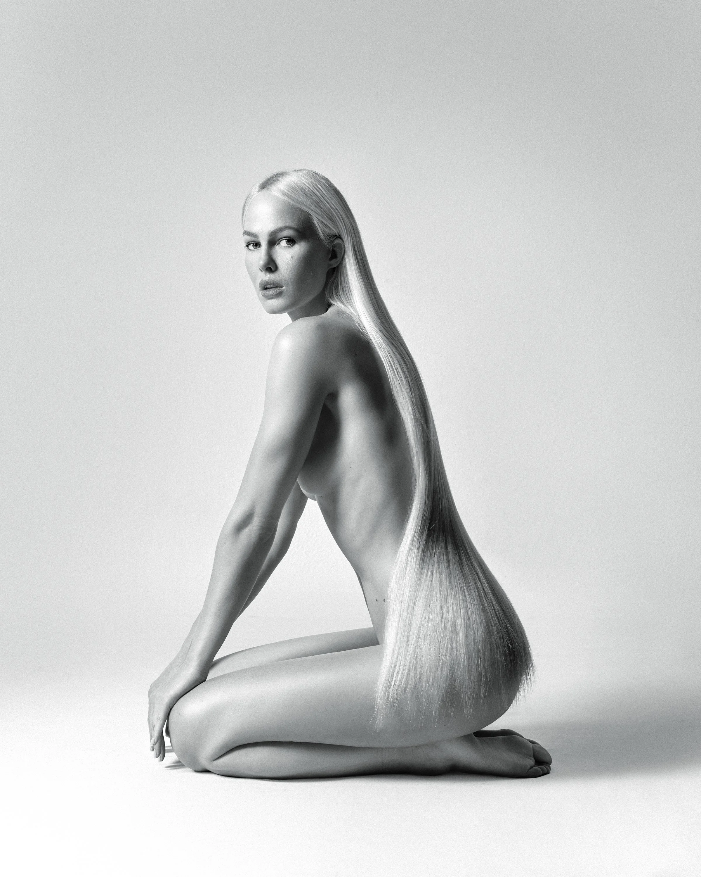 Black and white photo of a woman with long straight hair, sitting cross-legged on the floor, looking directly at the camera against a plain background.