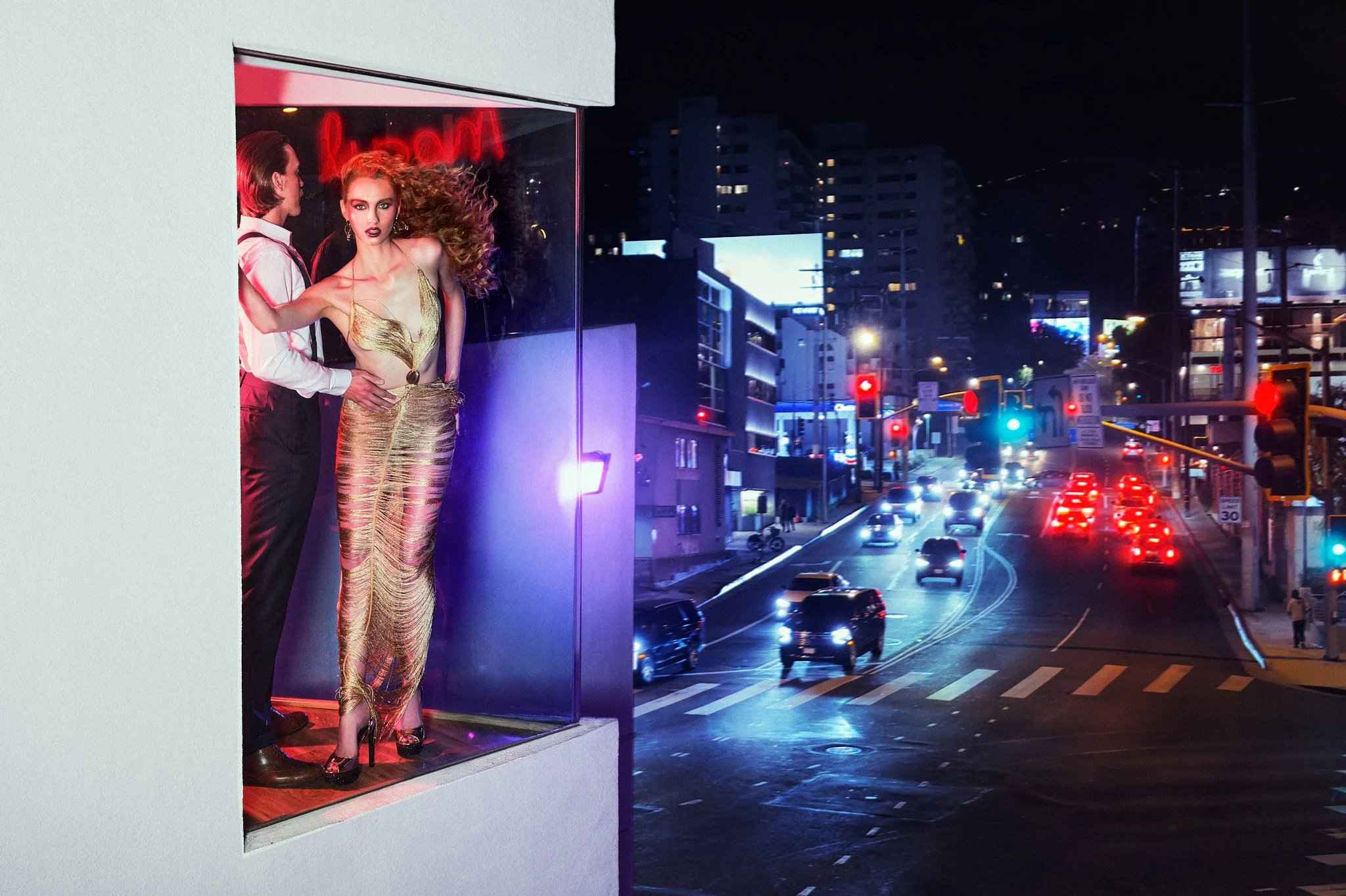 A woman in a shiny gold dress and high heels stands in a show window at night, with her hair blowing and a surprised expression. A man in a white shirt and sunglasses is partially visible behind her. The city street outside is busy with cars, traffic