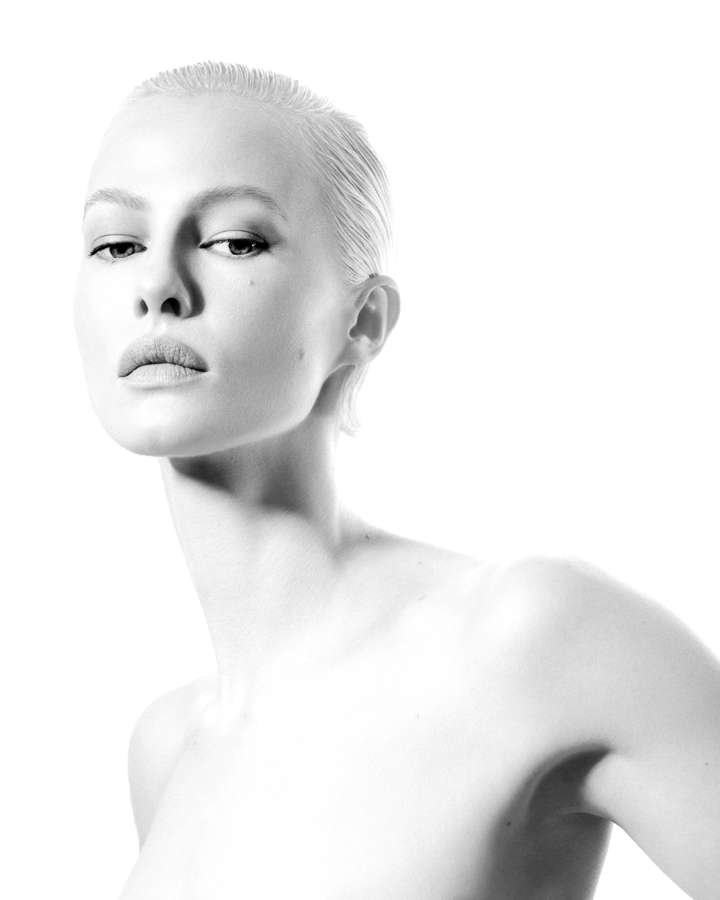 Close-up black and white portrait of a woman with platinum blonde hair slicked back, looking slightly to the side with a serious expression, minimal makeup, and bare shoulders.