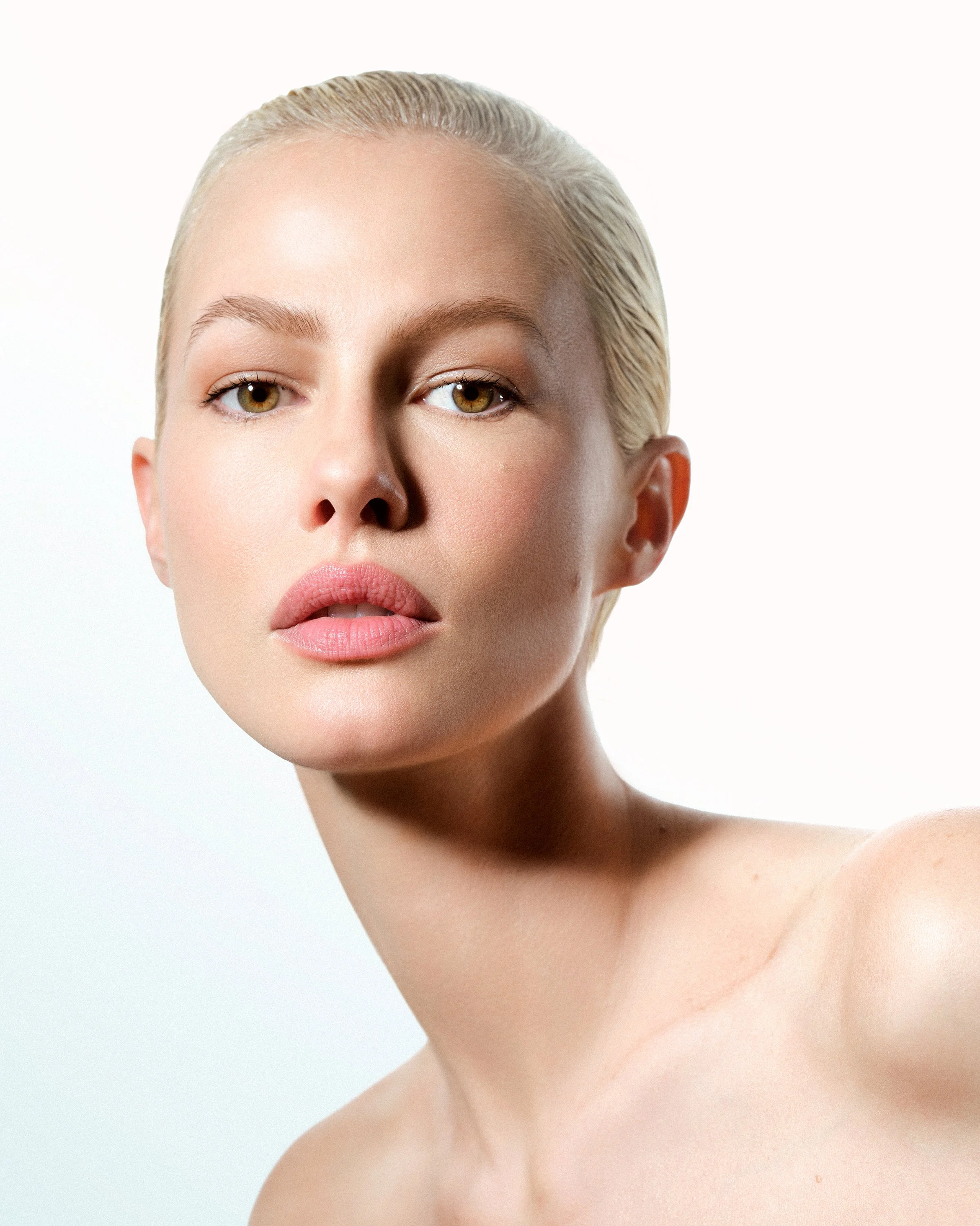 Close-up of a woman with blonde slicked-back hair, natural makeup, and bare shoulders against a white background.