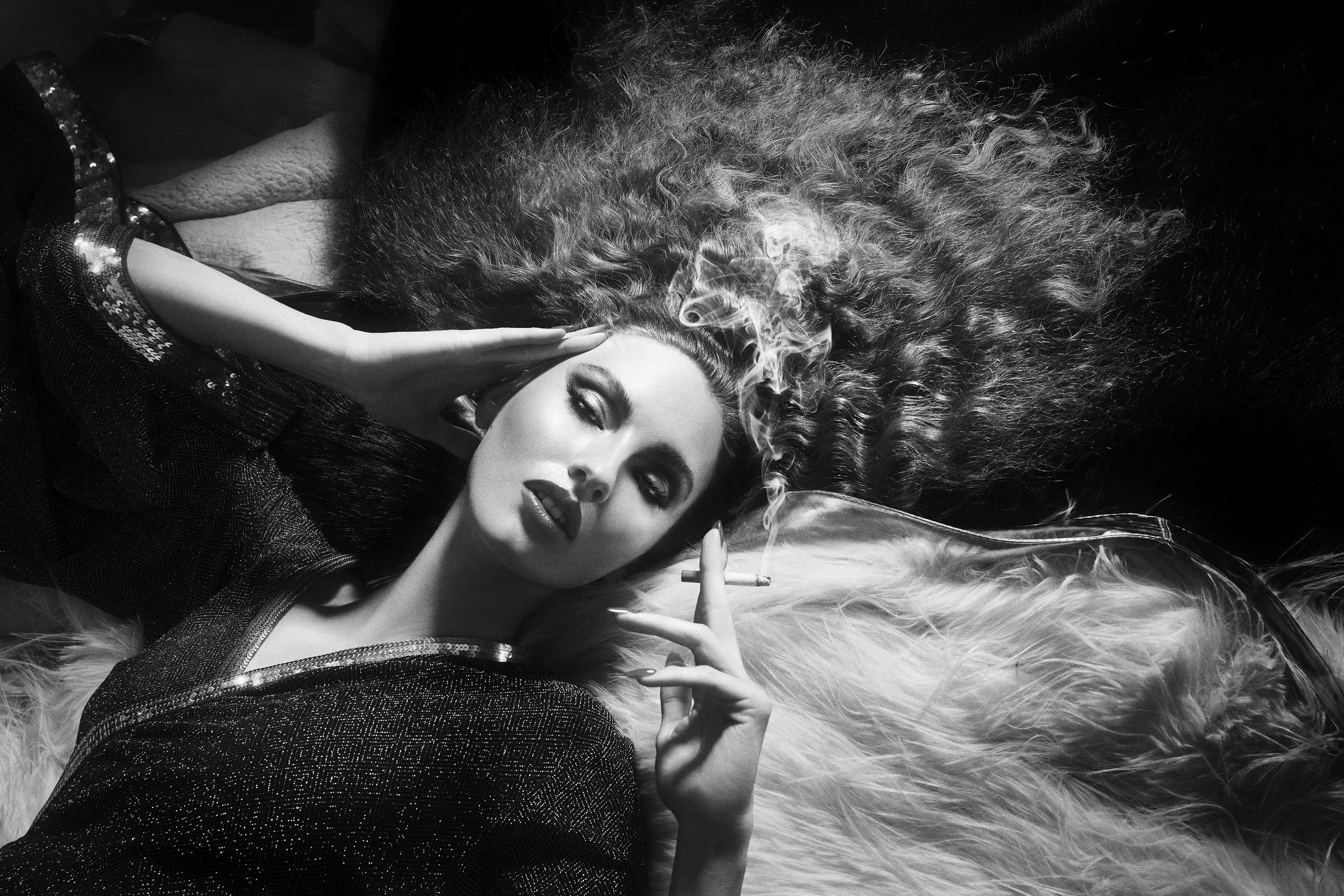 Black and white photo of a woman with curly hair smoking a cigarette, lying on a fluffy surface, with her eyes half-closed and a confident expression.