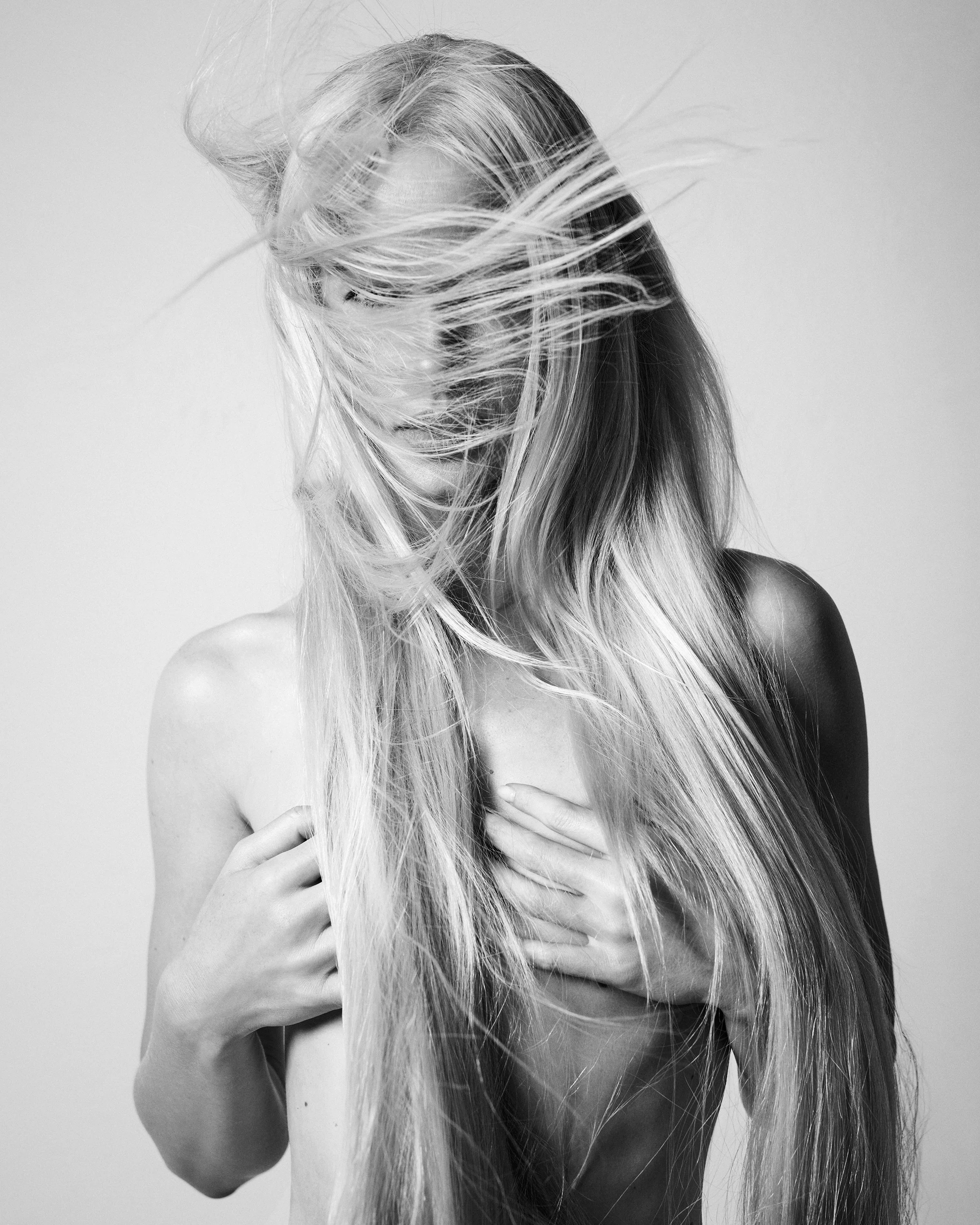 Black and white photo of a woman with long, flowing blonde hair covering her face, with her hands crossing her chest.
