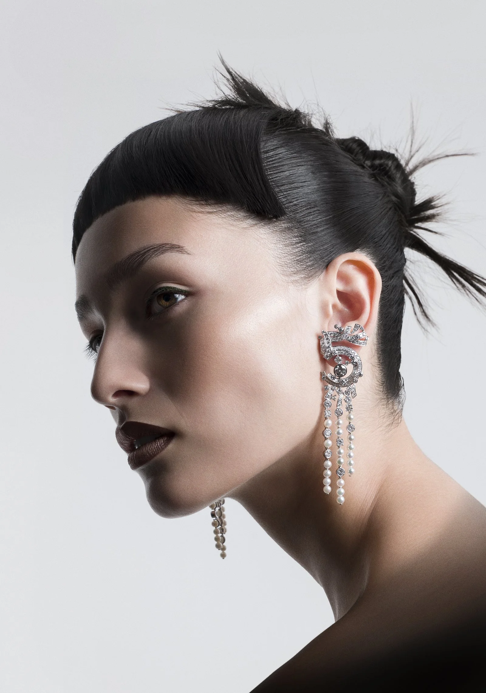Close-up of a woman's face with dark, styled hair, wearing elaborate pearl and rhinestone earrings.