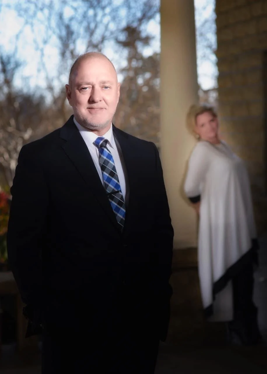 A man in a black suit and a blue plaid tie standing outside near brown brick columns, with a blurred woman in a white cape and black dress in the background.