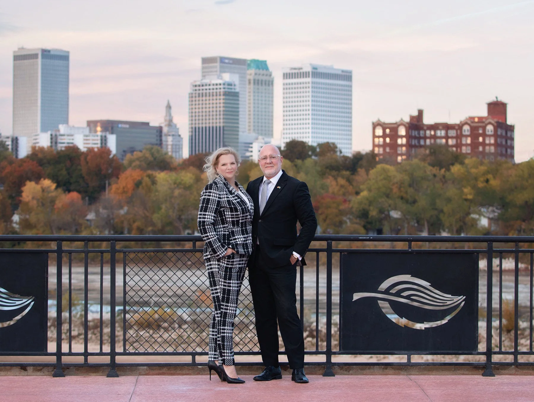 A man and a woman standing side by side on a balcony with a city skyline and trees with autumn foliage in the background
