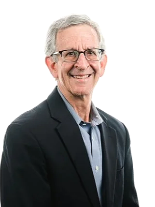 A smiling older man with gray hair, glasses, wearing a black blazer and light blue shirt, posing against a plain white background.
