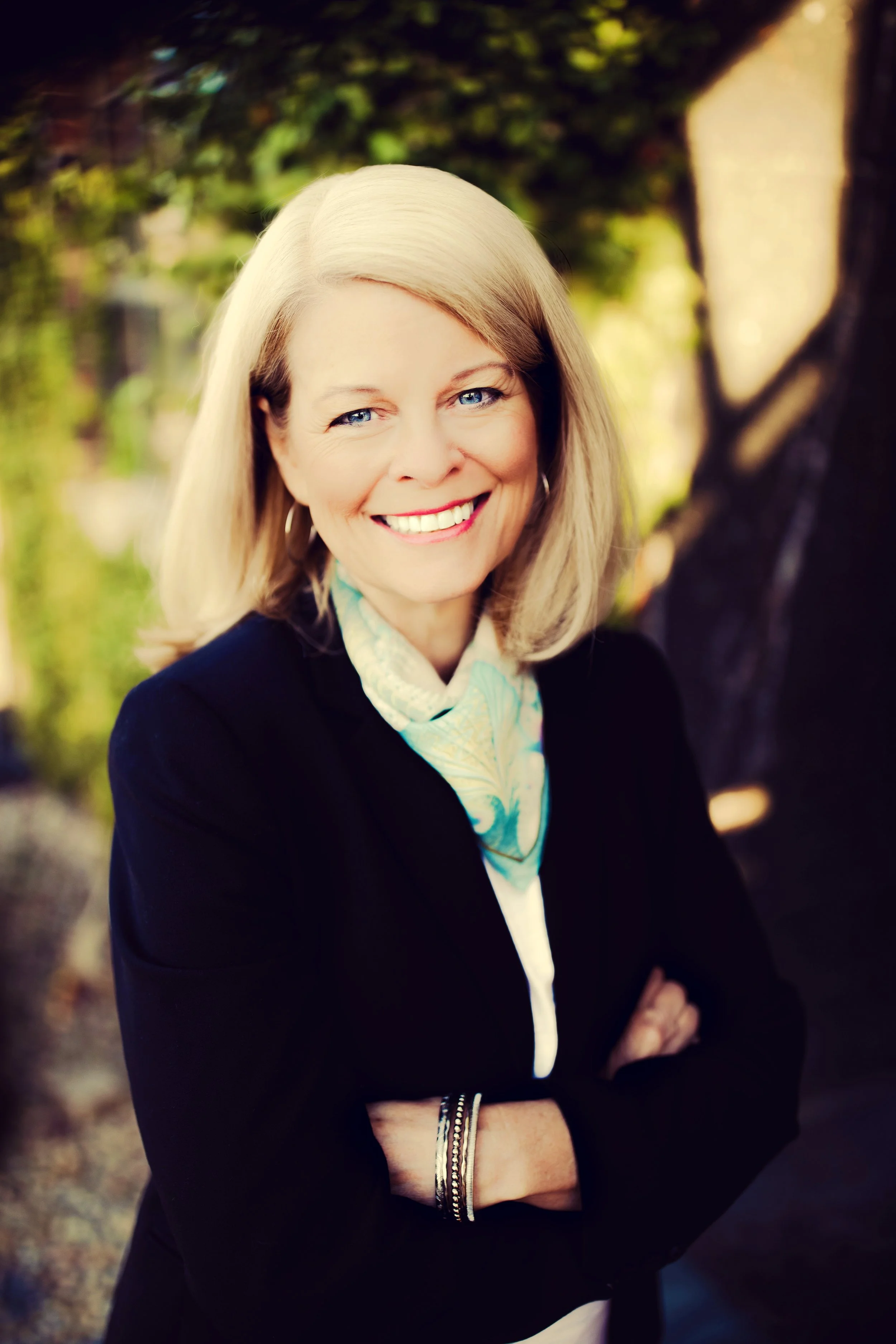 A woman with blonde hair, smiling, wearing a black blazer, a light-colored scarf, and bracelets on her wrist, standing outdoors with blurred trees and greenery in the background.