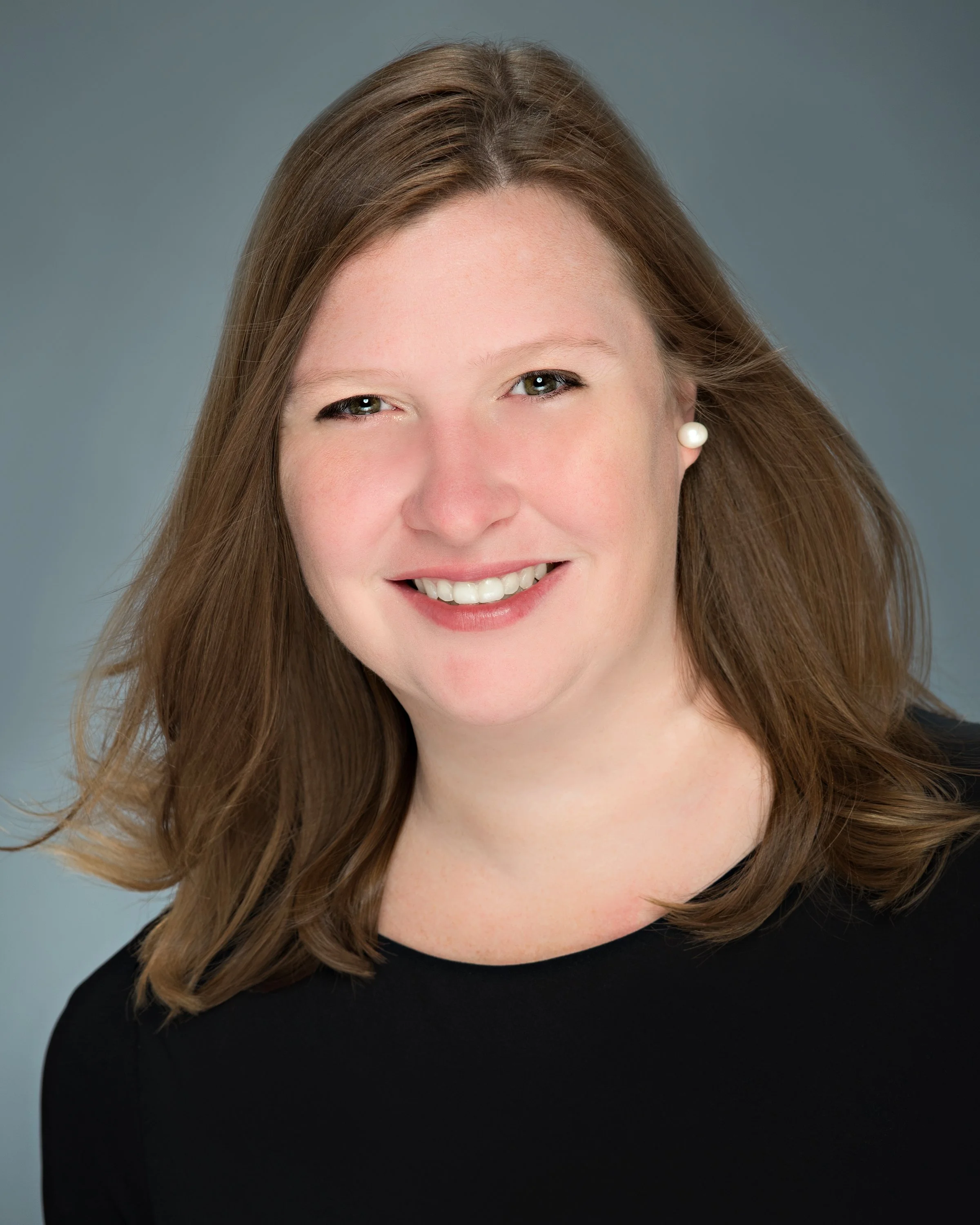 Headshot of a woman with shoulder-length brown hair, smiling, wearing pearl earrings and a black top, against a gray background.