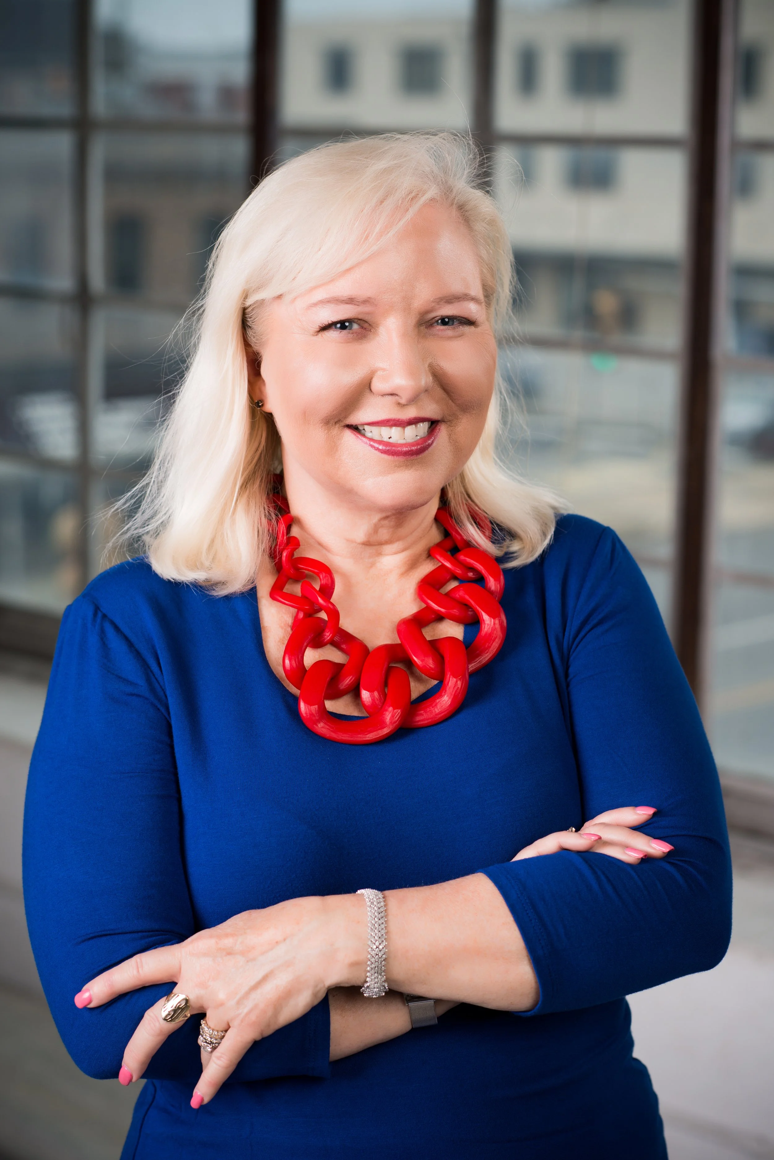 Portrait of a smiling woman with blonde hair wearing a blue dress and a chunky red necklace, standing in front of large windows.