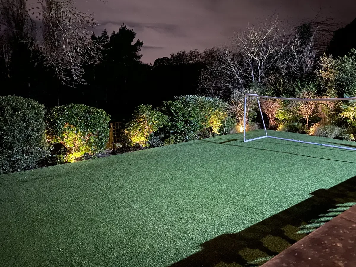 Back garden at night with strategically placed uplights enhancing bushes and trees, adding elegance and depth to the landscape through well-designed outdoor lighting.