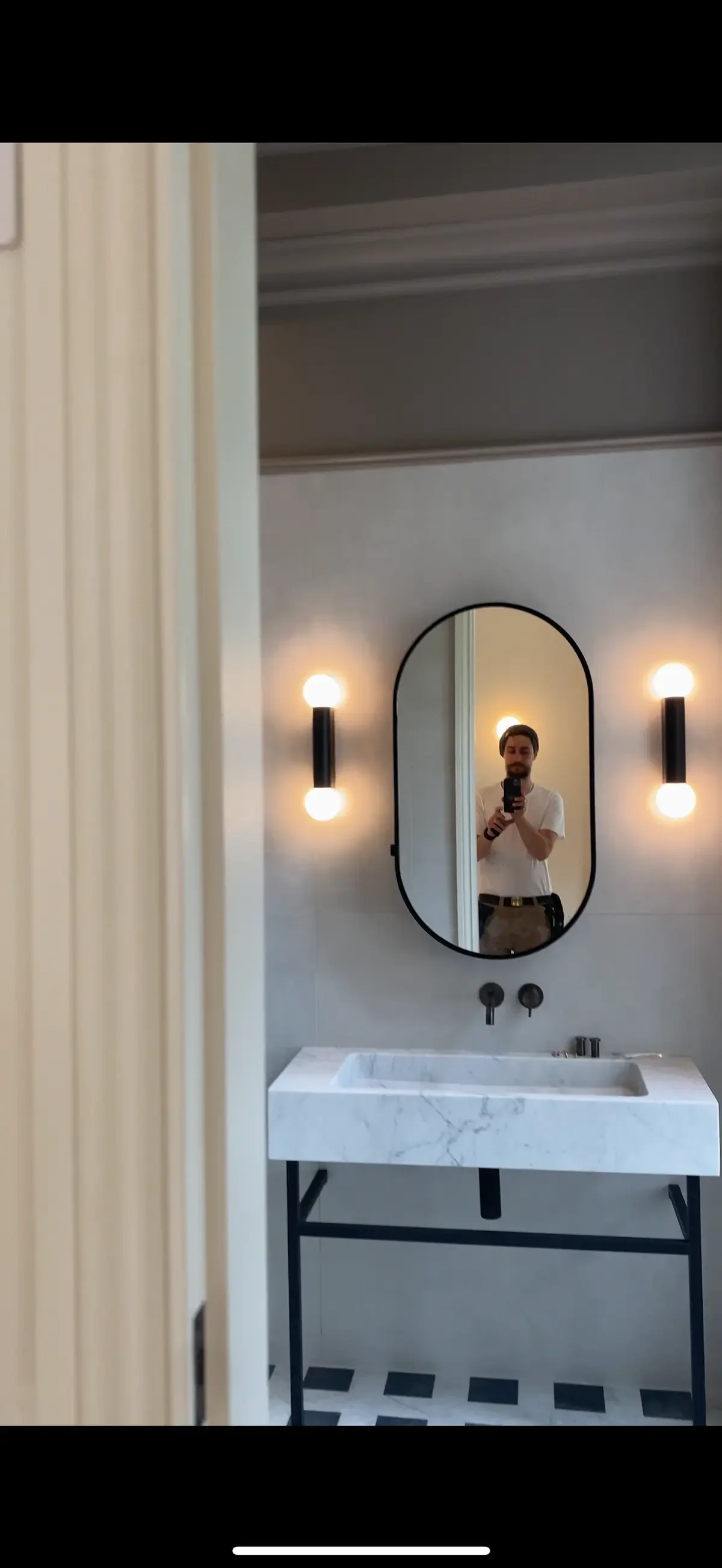 Contemporary bathroom with black wall lights flanking an oval mirror above a marble sink, showcasing clean electrical installations and ambient lighting design.