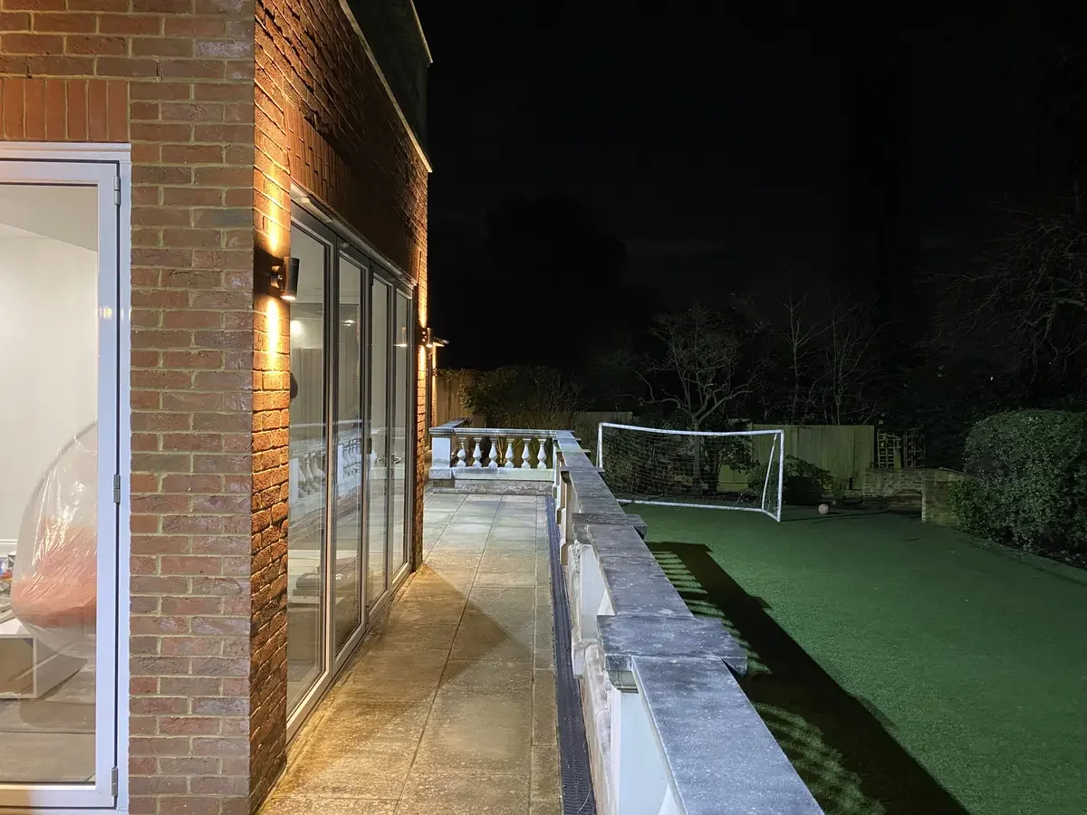 Outdoor scene of a brick home with warm wall-mounted exterior lights illuminating a garden terrace and artificial turf area with a football goal at night.