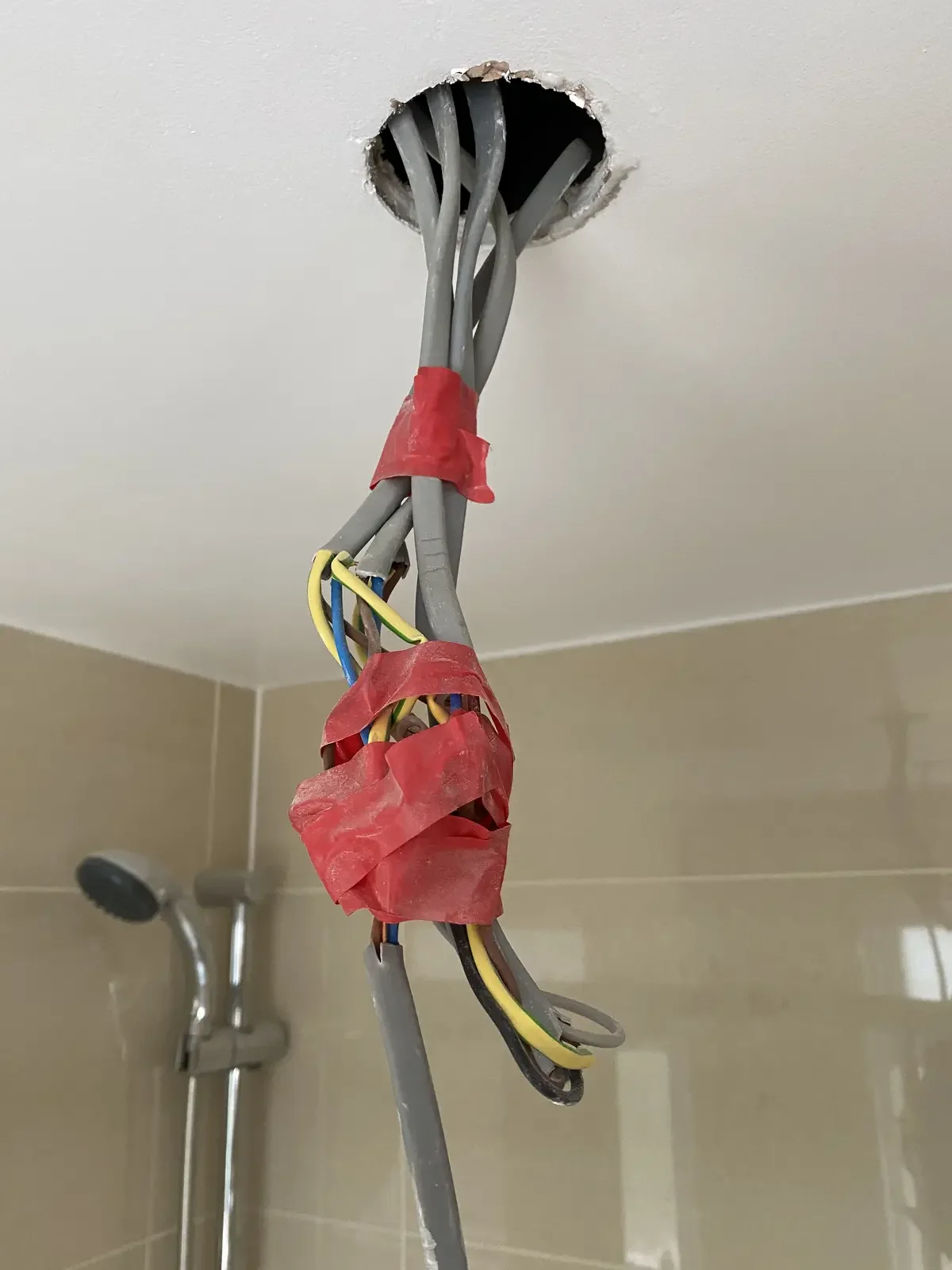 Multiple electrical wires wrapped with red electrical tape hanging from a ceiling, with a shower area visible in the background.