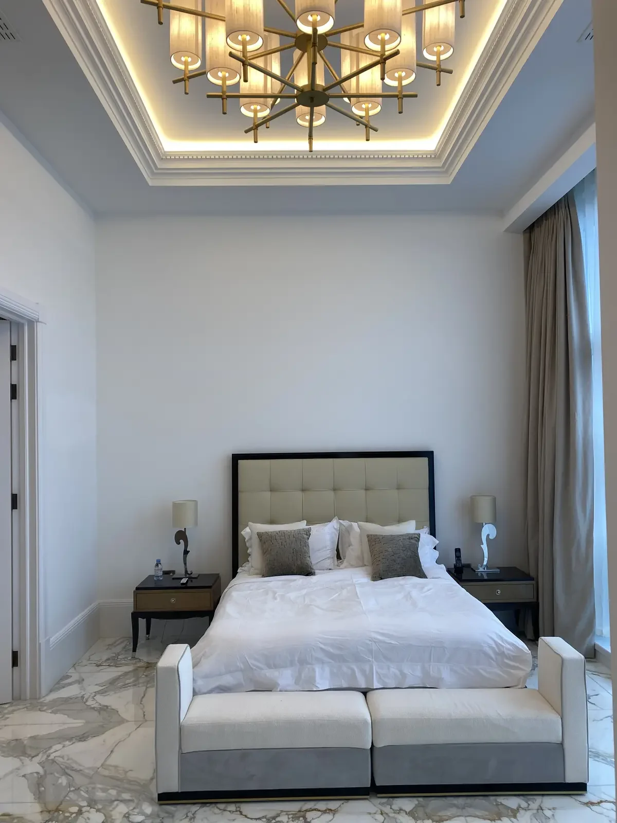 Spacious master bedroom with inset ceiling lighting and a multi-arm chandelier, framed by elegant floor-to-ceiling curtains and marble tile flooring.