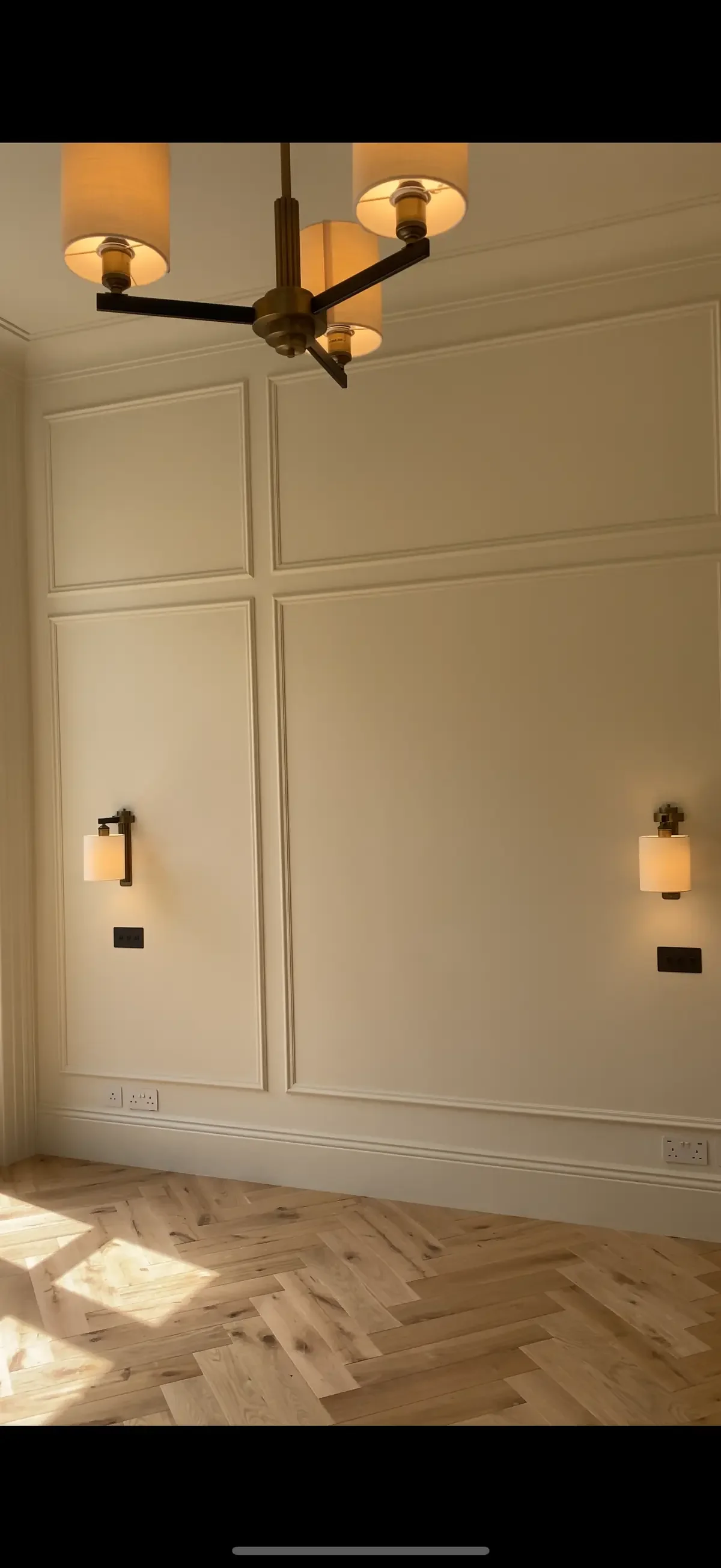 Elegant cream-colored wall with panel mouldings featuring symmetrical warm wall sconces and a central chandelier, highlighting premium interior lighting and electrical finish.