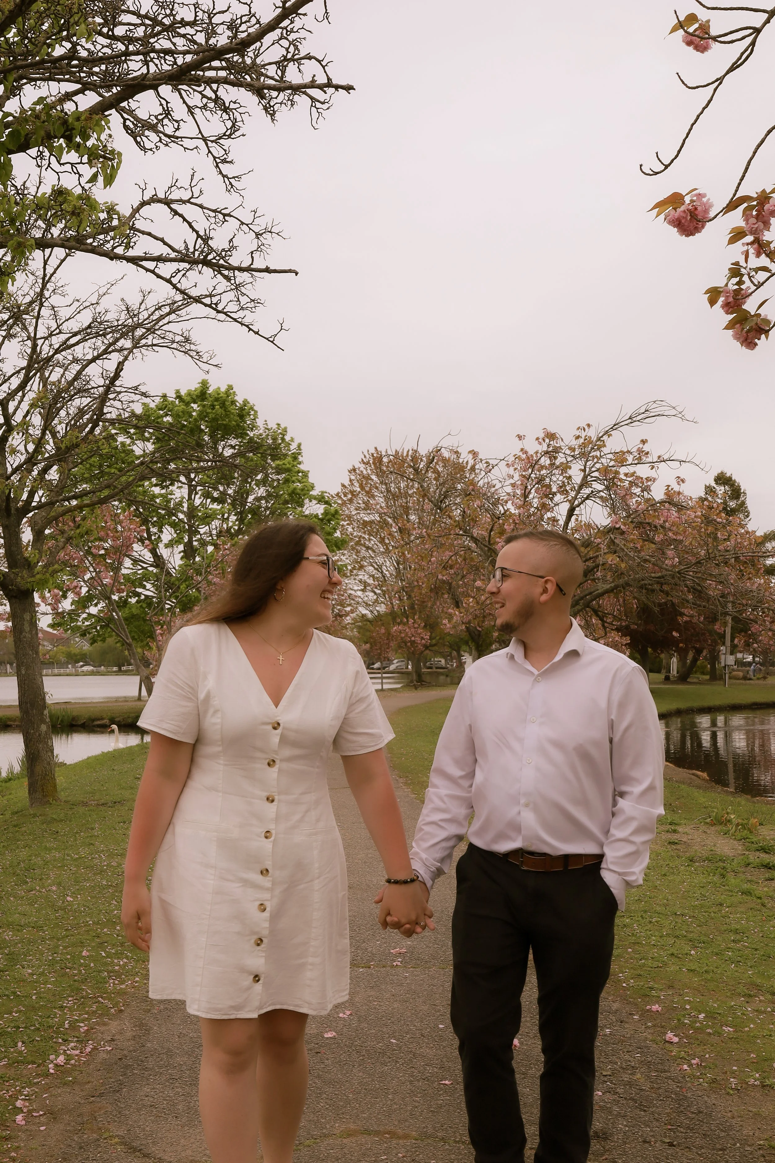 A couple walking hand-in-hand in a park with pink blossoms and trees, smiling at each other.