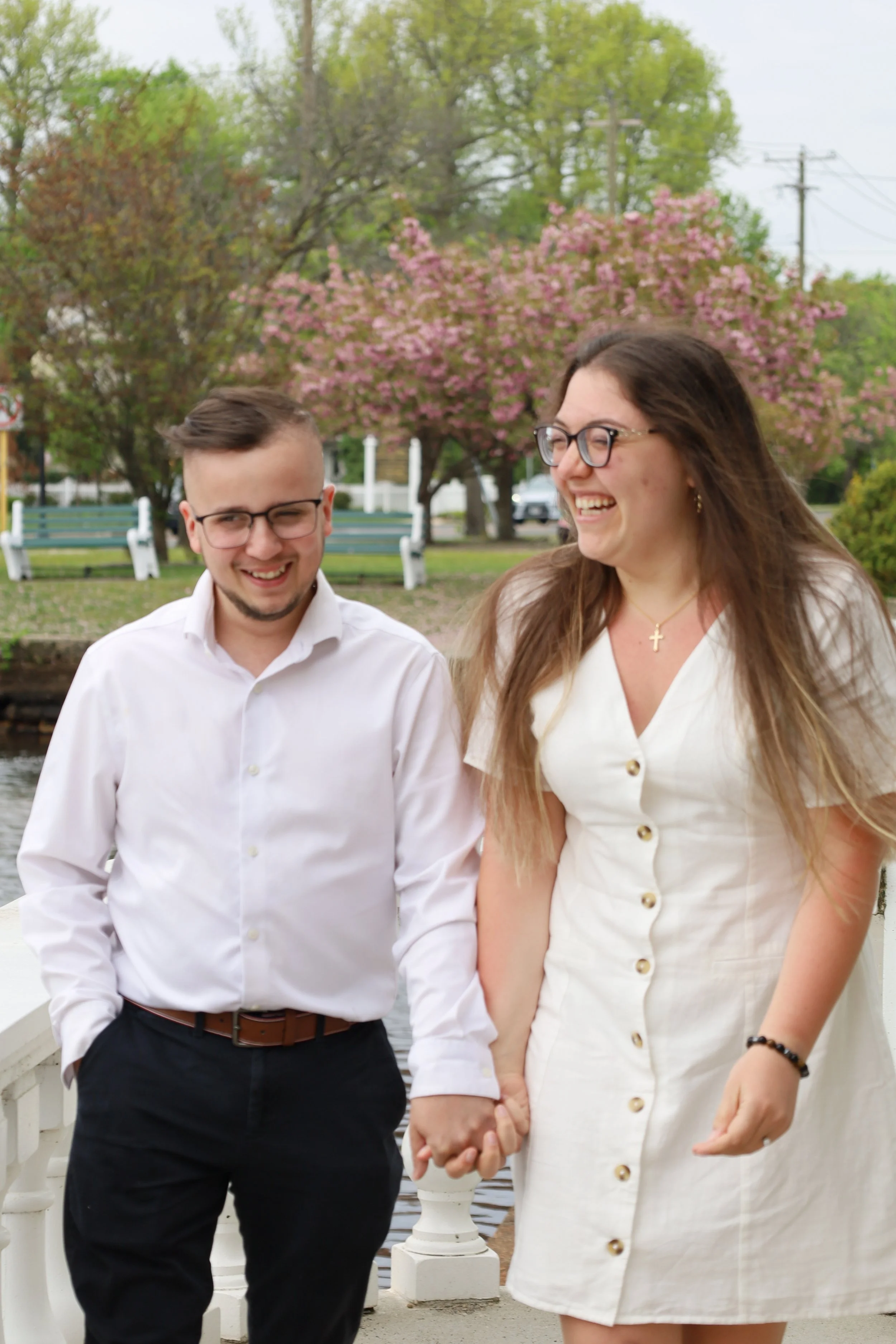 A man and woman holding hands, smiling, walking outdoors with trees and pink blossoms in the background.