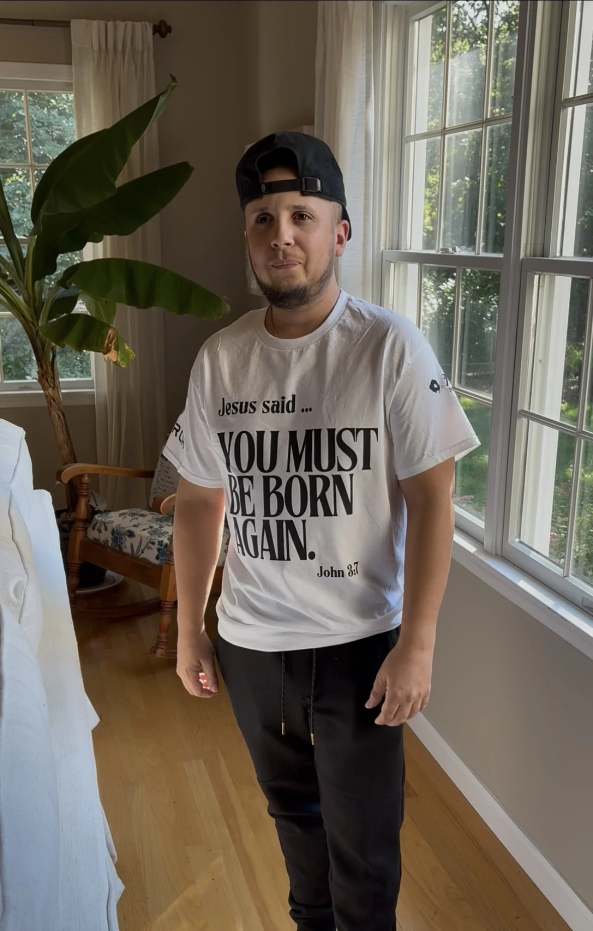 A man wearing a white T-shirt with a Bible quote from John 3:7