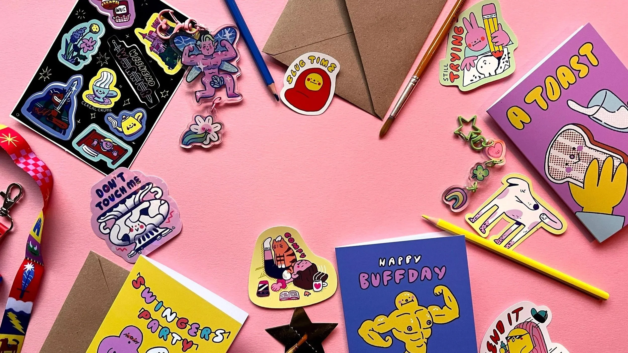 A flat lay of colorful stickers, stationery, and decorative items on a pink background. Includes stickers of animals, characters, and humorous phrases, along with pens, envelopes, and a lanyard.