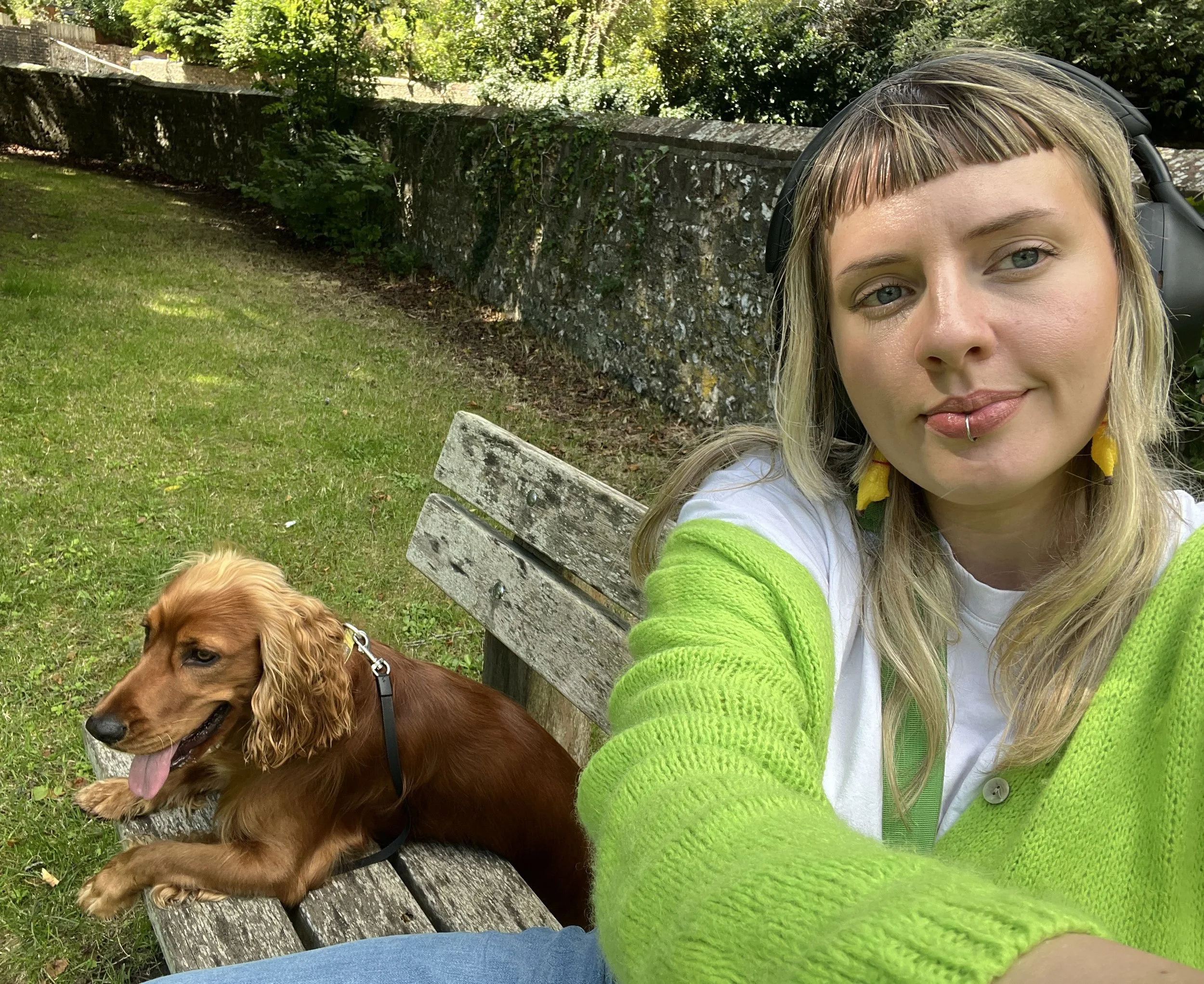 A woman with blond hair, wearing a white shirt and bright green sweater, taking a selfie outdoors on a park bench. She has a lip piercing and is wearing yellow earrings. Next to her on the bench, a brown dog with floppy ears, lying down with tongue out, wearing a collar.