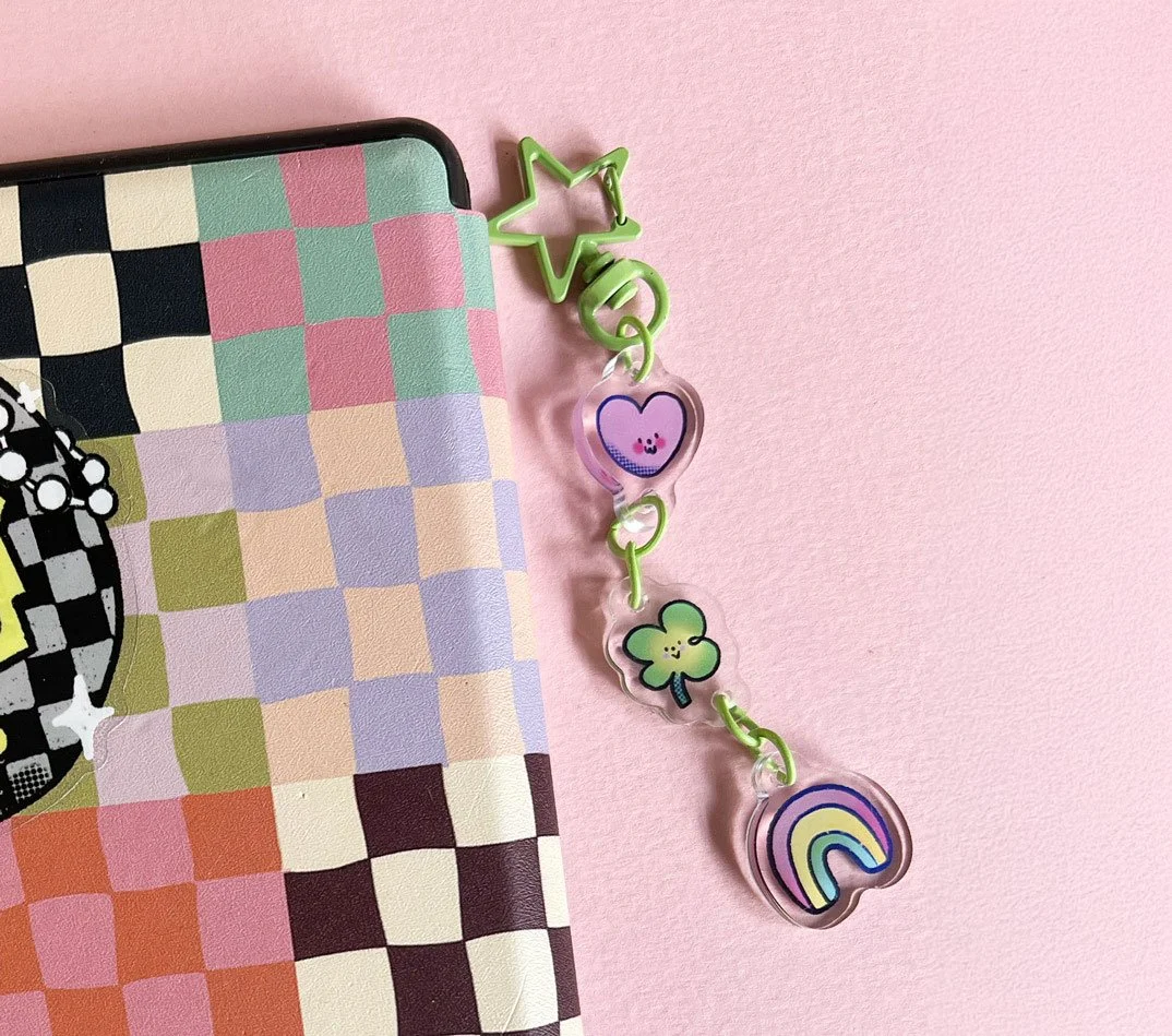 Colorful rainbow, shamrock, pink heart, and star-shaped keychain attached to a green clip on a pink background next to a notebook with a checkered pattern.