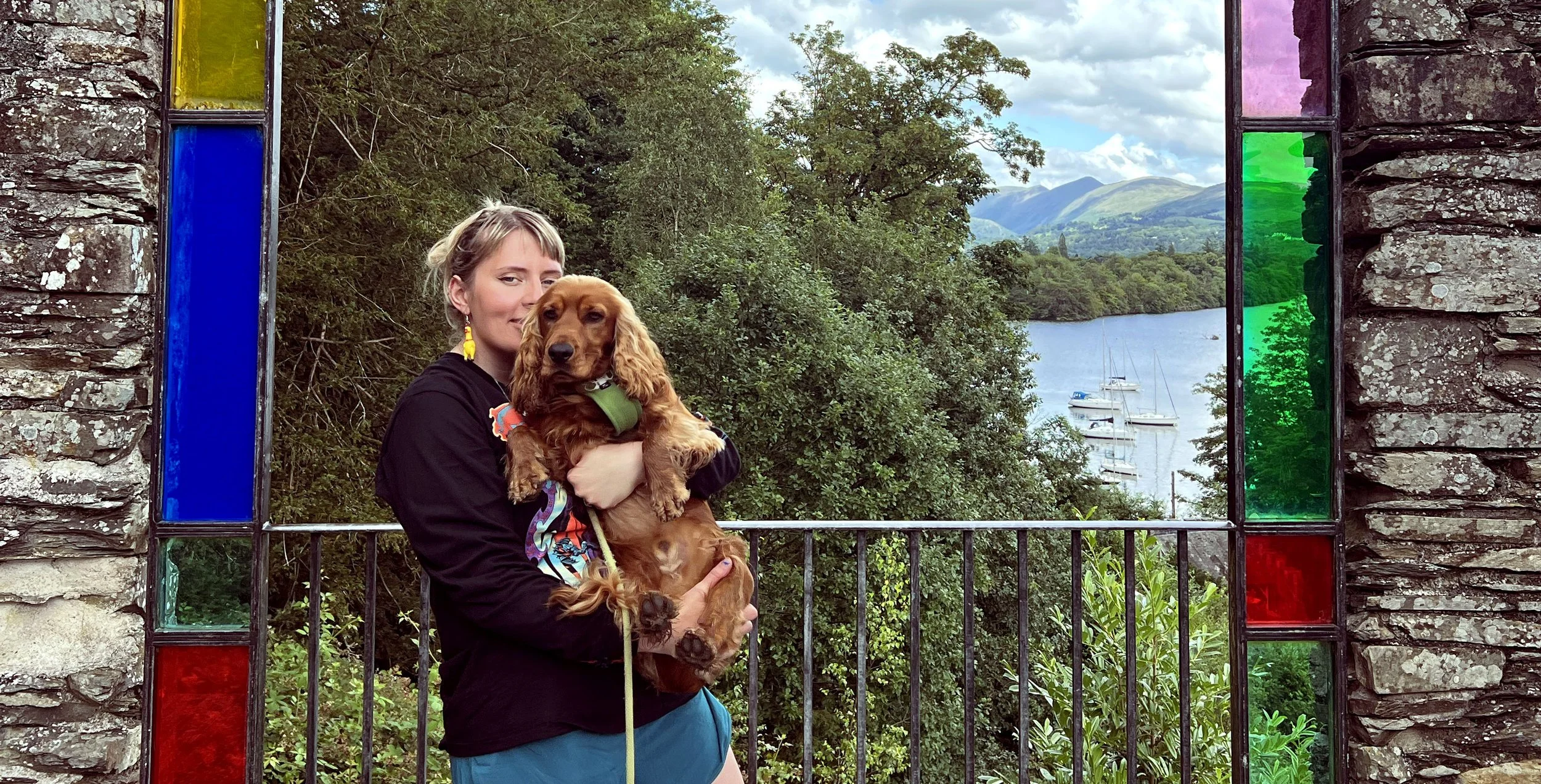 A woman holding a dog in front of a stained glass window with a scenic view of trees, mountains, and boats on a lake.