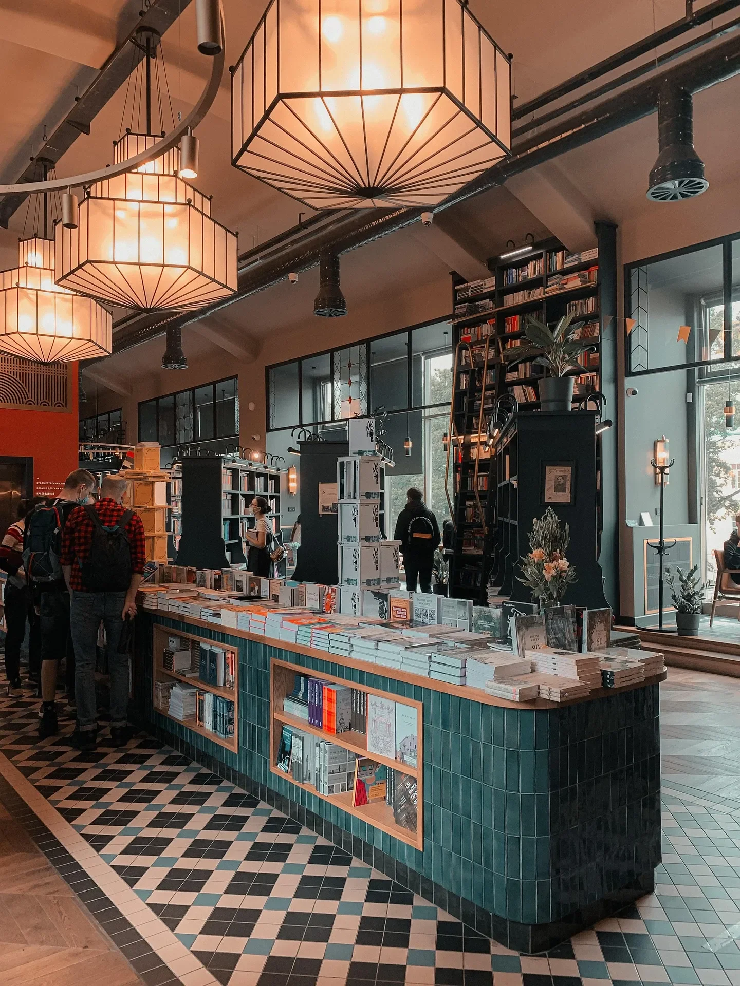 The inside of a small bookstore with a book display; people browsing; modern, hexagonal lighting fixtures; and large windows.