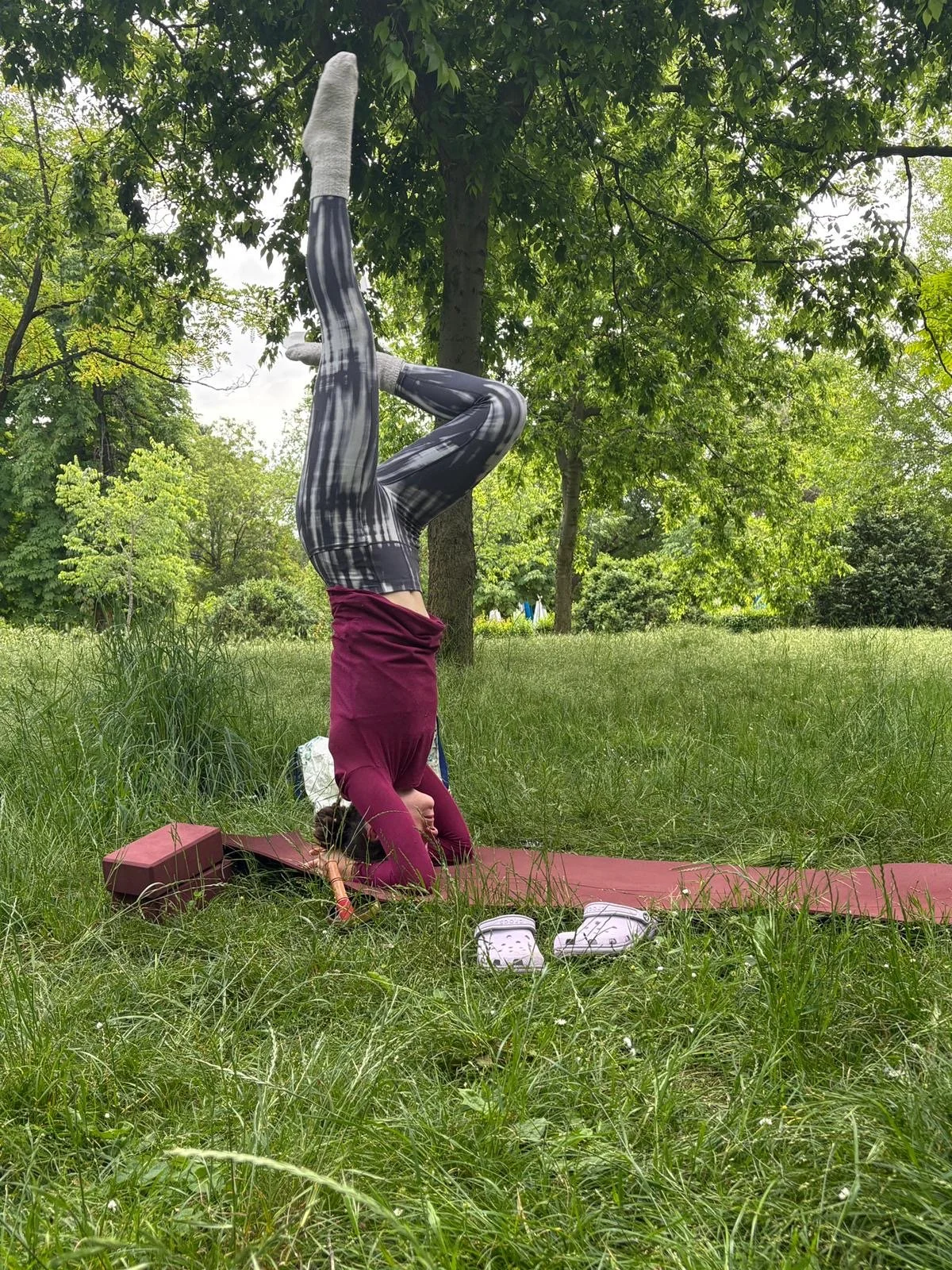 A person doing a headstand yoga pose outdoors on a grassy field, with trees and green foliage in the background.