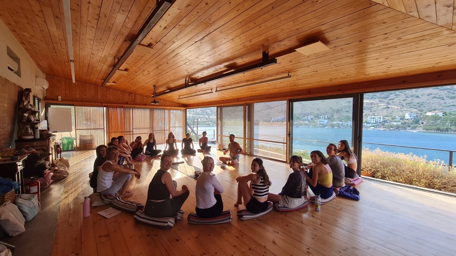 Group of people sitting cross-legged on yoga mats in a spacious, wood-floored room with large windows overlooking a lake and mountains.