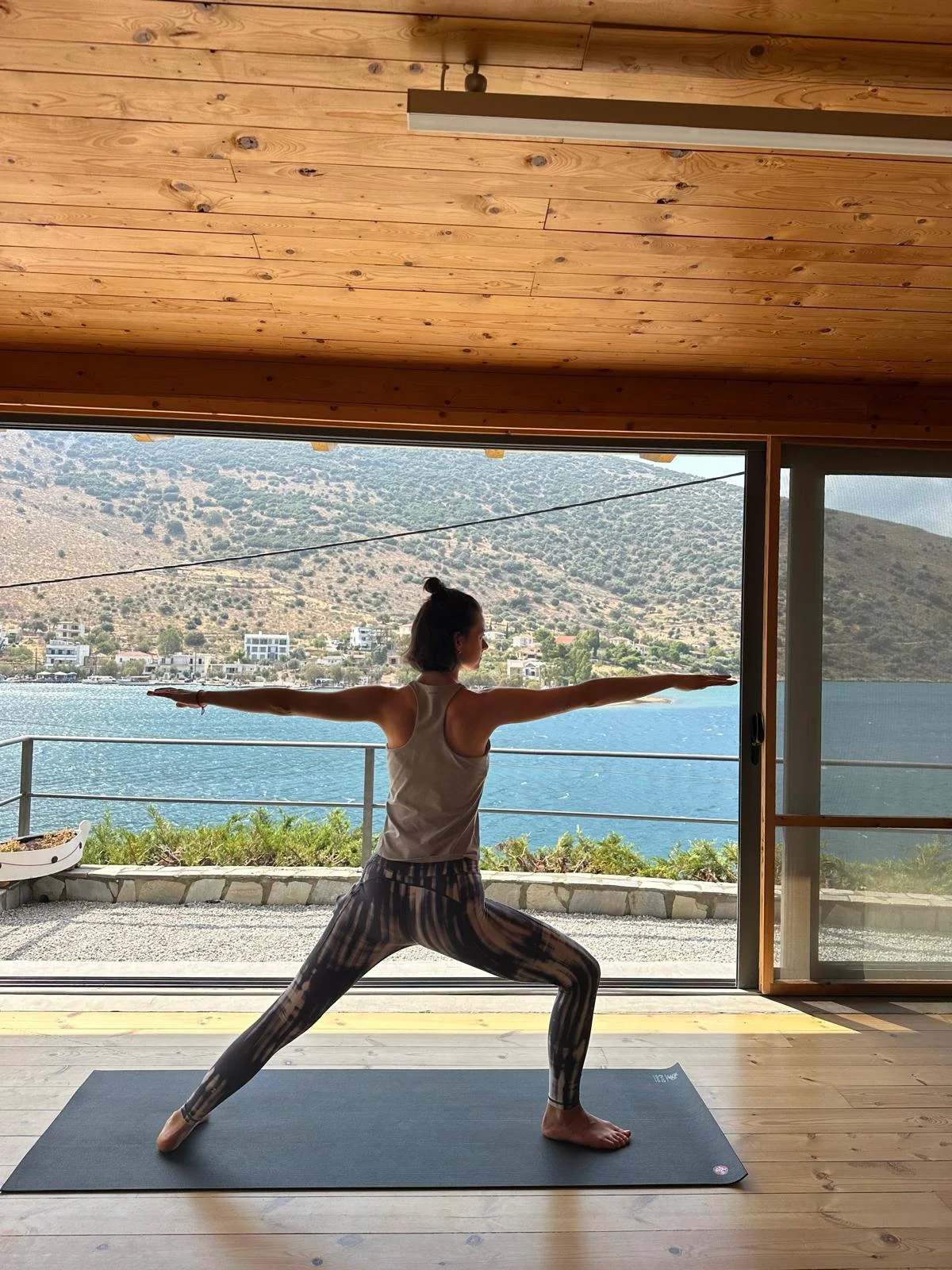 A woman practicing yoga indoors on a black mat with a scenic view of water, houses, and hills through large windows. She is in a Warrior pose with arms extended and legs wide apart.