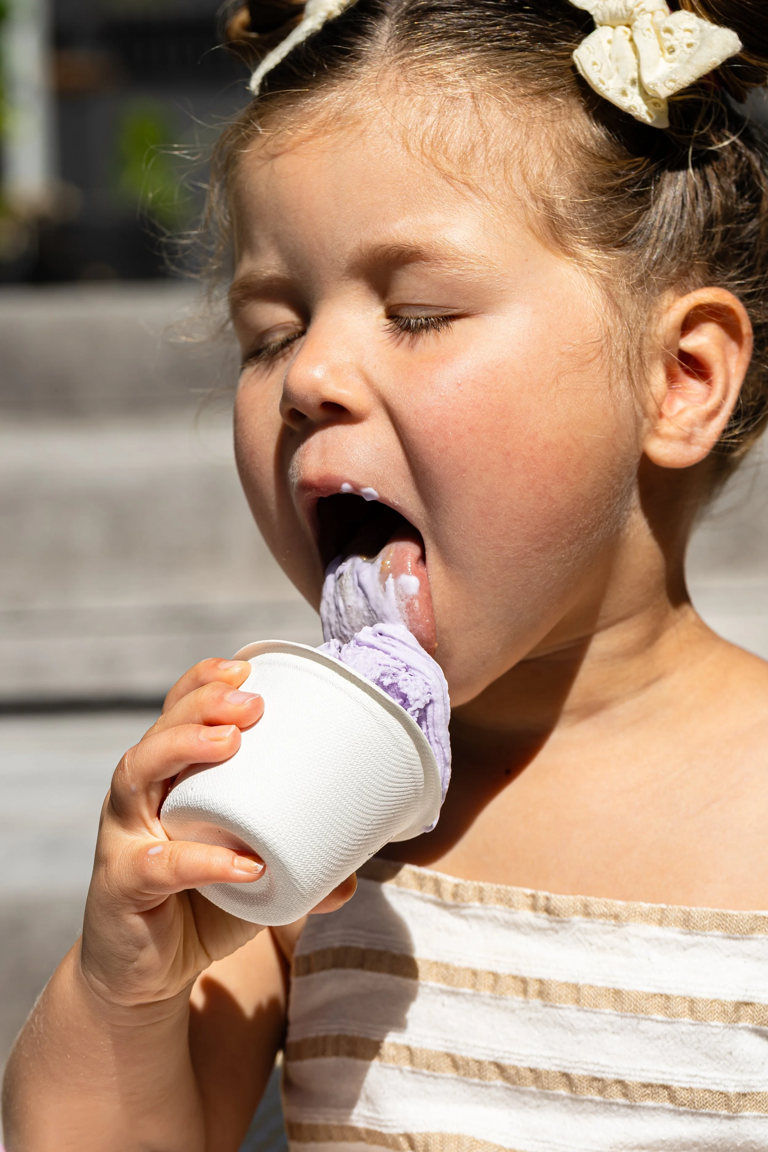A young girl with curly hair tied with a bow is enjoying a scoop of purple ice cream outdoors.