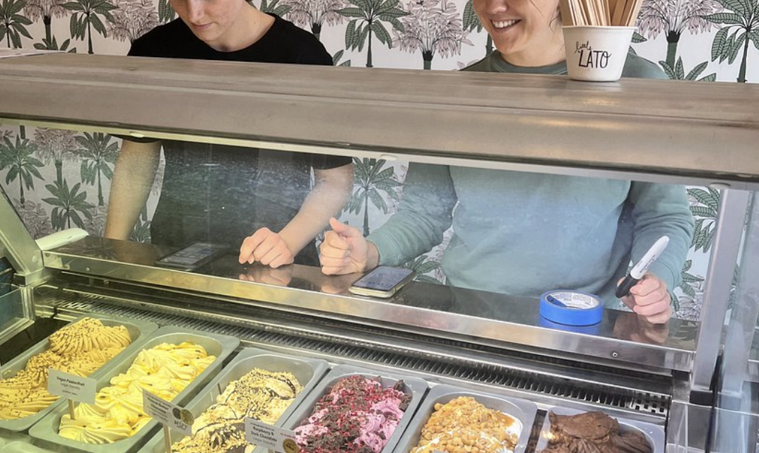 People selecting ice cream at a glass display case with various flavors, including yellow, white, and pink, in a shop with tropical-themed wallpaper.