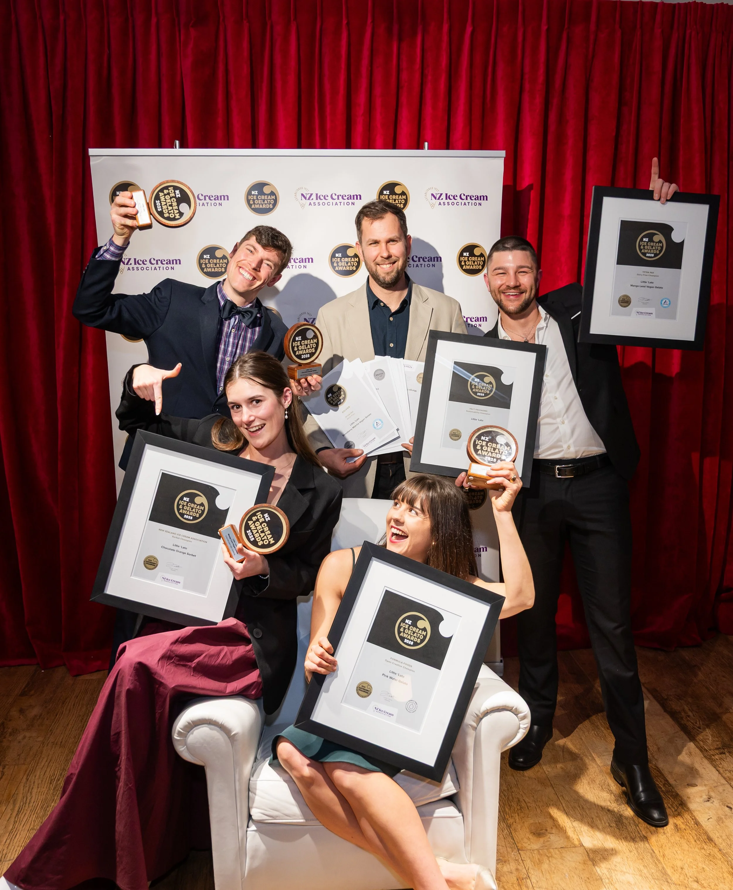 Group of people at an awards ceremony, holding framed certificates and trophies, standing in front of a backdrop with logos, and red curtains in the background.