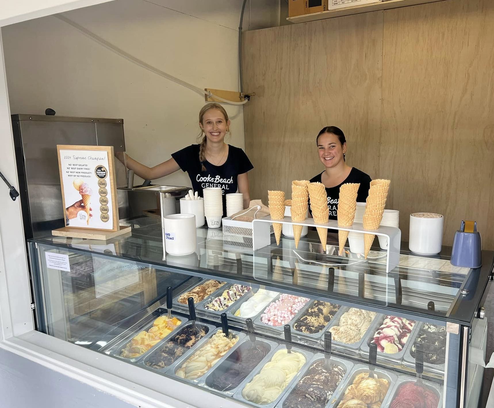 Two women stand behind an ice cream stand with multiple flavors of ice cream and cones on display. One woman is smiling and posing, while the other is smiling and looking at the camera. The stand has a sign with ice cream options and a small display of ice cream cones.