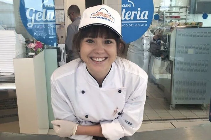 Smiling female ice cream shop worker wearing a white uniform and cap, standing in front of a gelato shop counter.