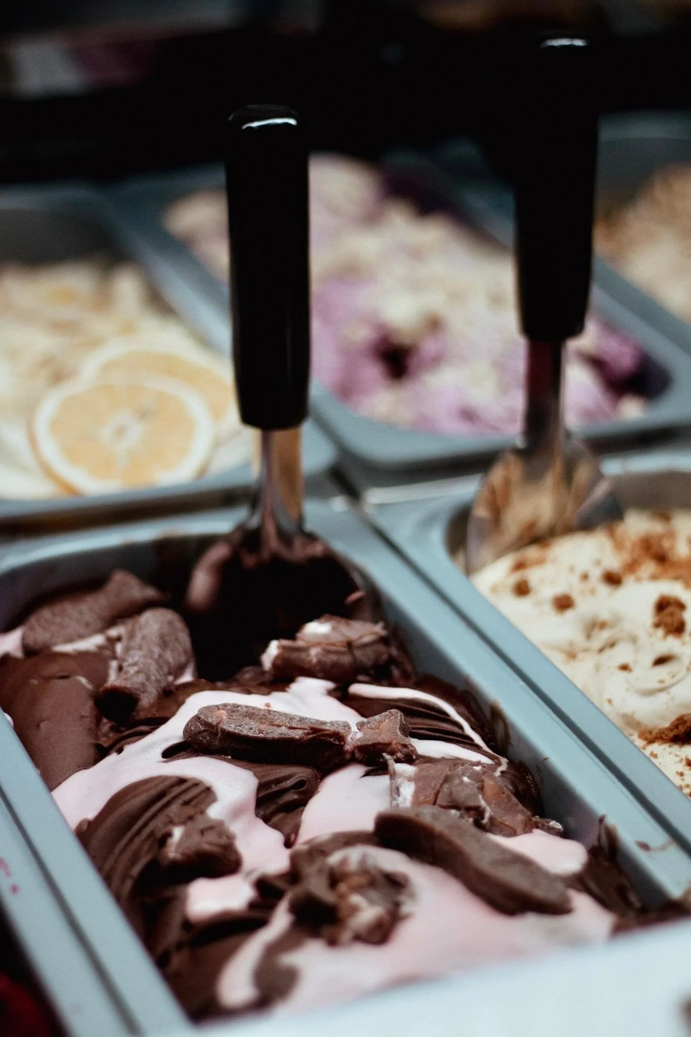 Close-up of multiple tubs of different flavors of ice cream, including chocolate, vanilla, and berry, in a freezer display, with ice cream scoops resting on top.