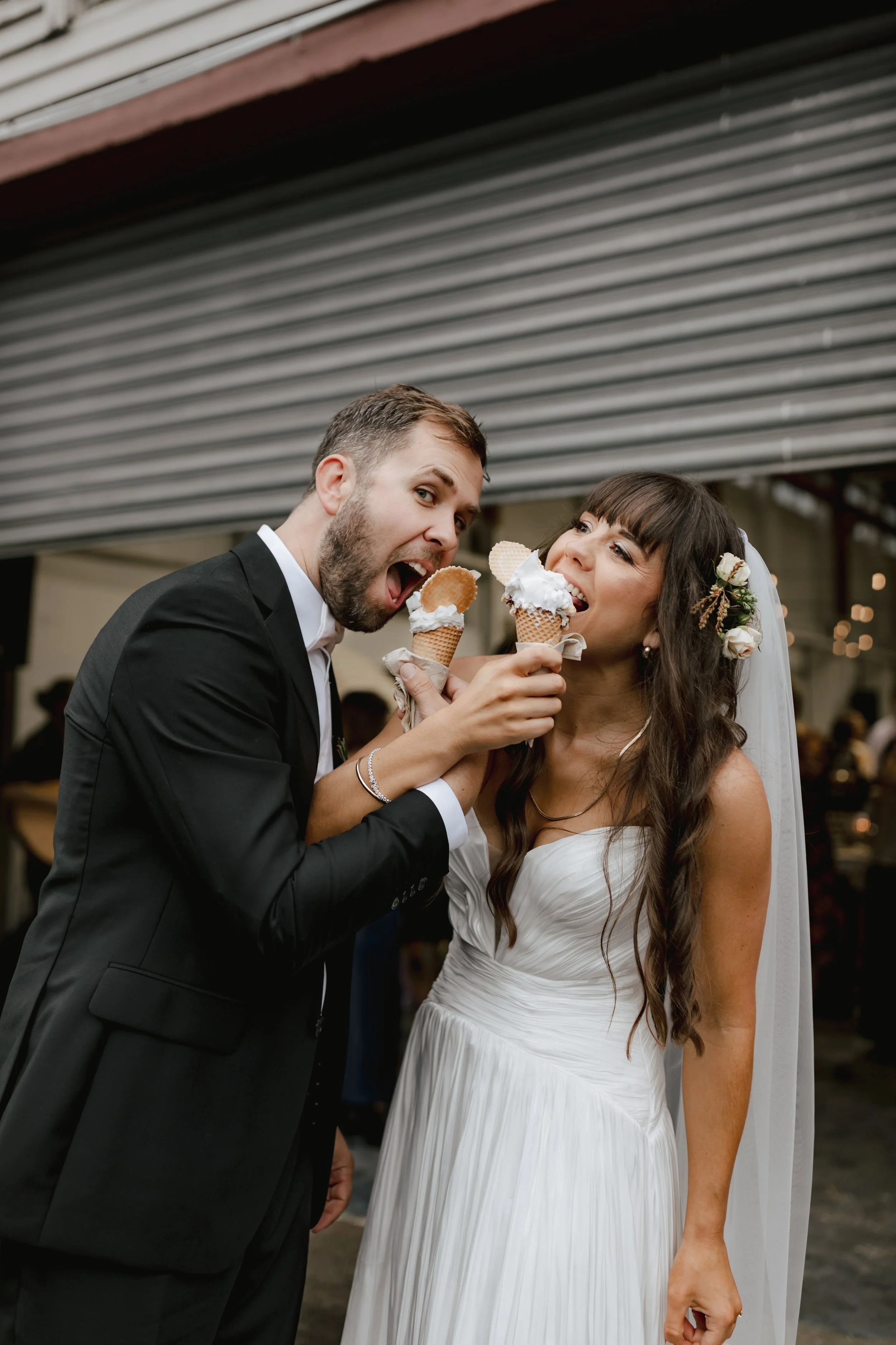 A man and a woman, dressed in wedding attire, standing close together at a wedding reception, happily eating ice cream cones.