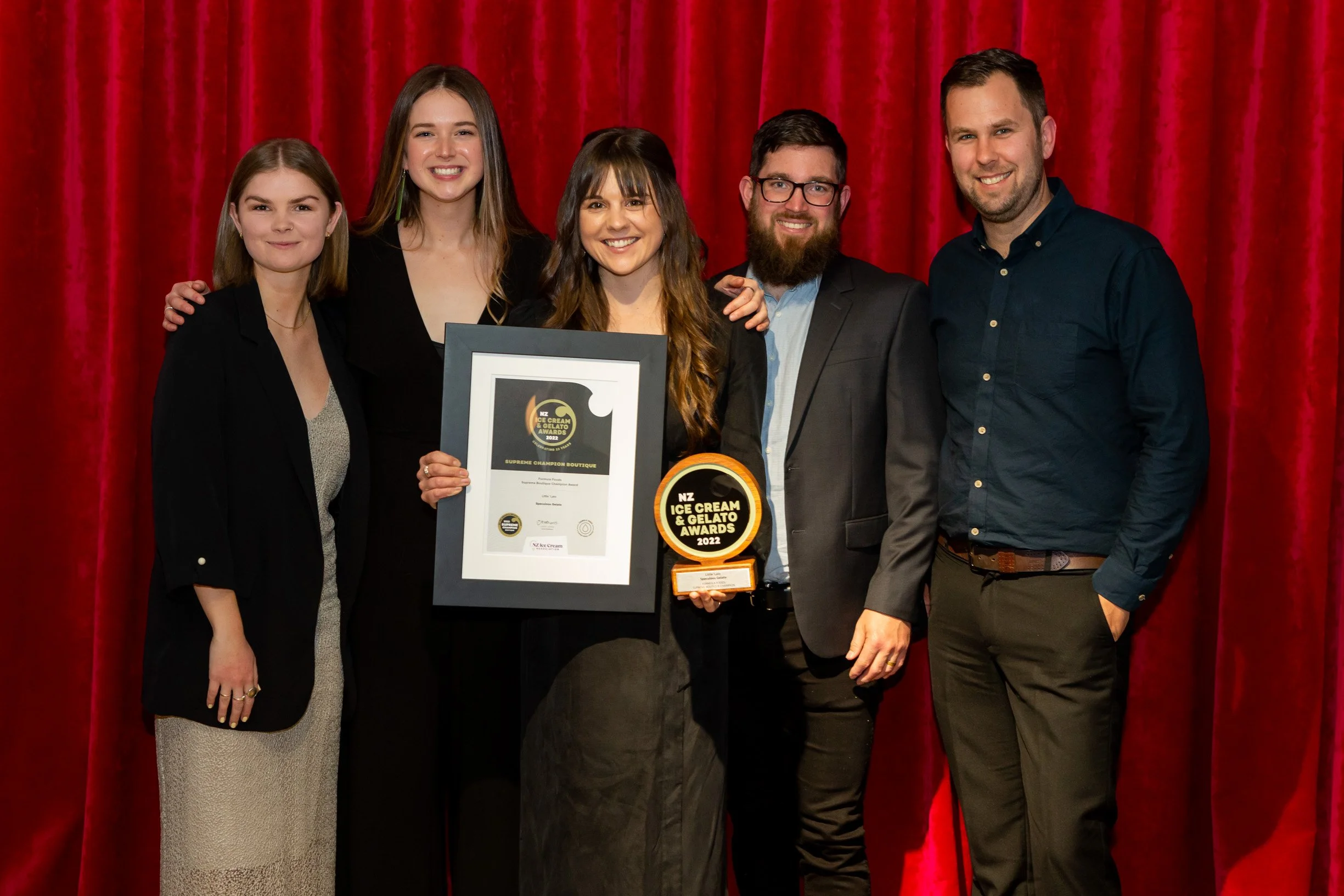 Group of six people, three women and three men, standing together in front of a red curtain at an awards event. They are smiling, with one holding an award plaque and another holding a framed certificate.
