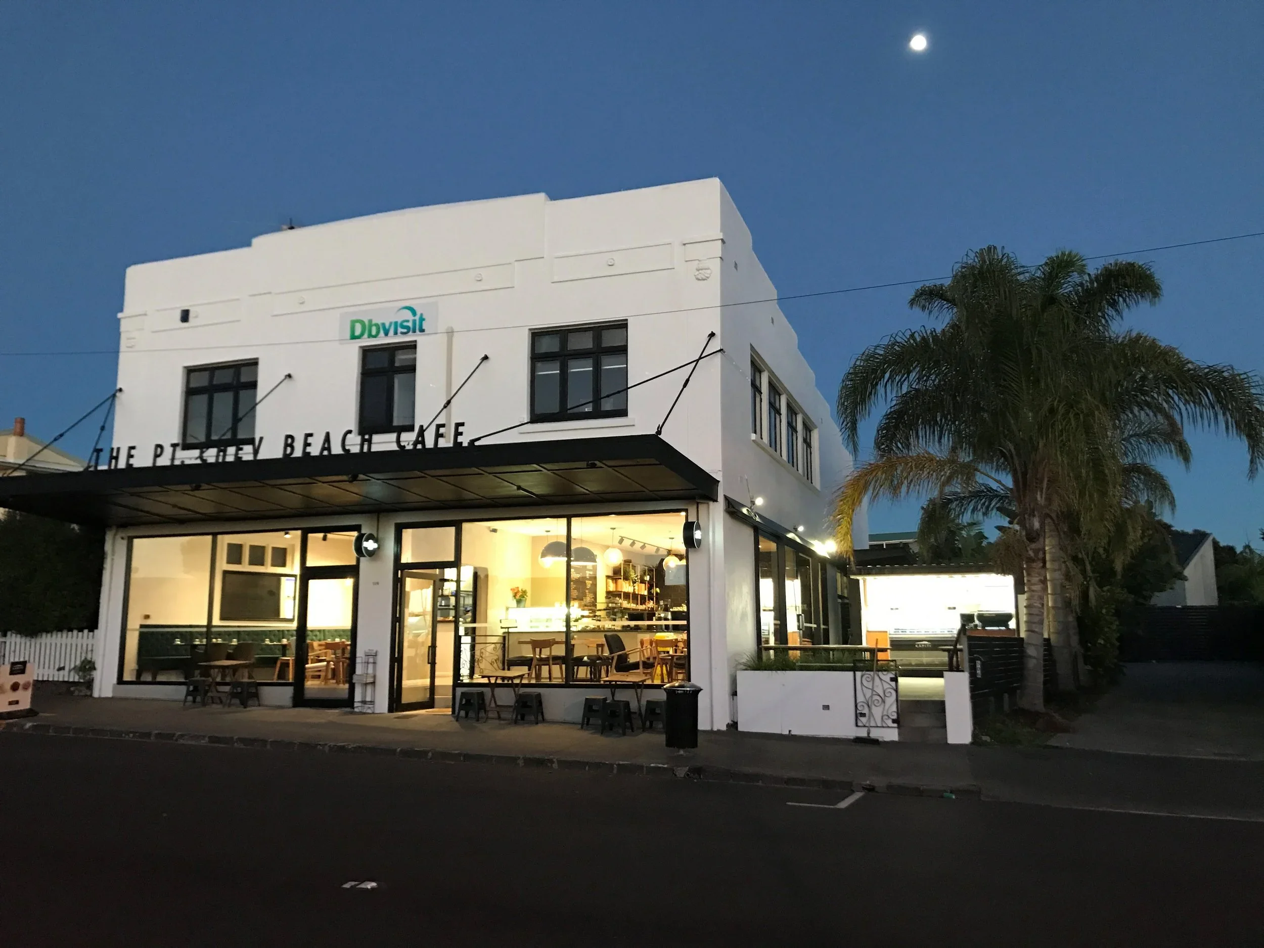 A two-story white building with black-framed windows and a sign that reads 'The Pollen Beach Cafe' at night. The building has a well-lit interior visible through large glass windows, with outdoor seating in front. The sky is dark with a visible bright moon, and there is a tall palm tree on the right side of the building.