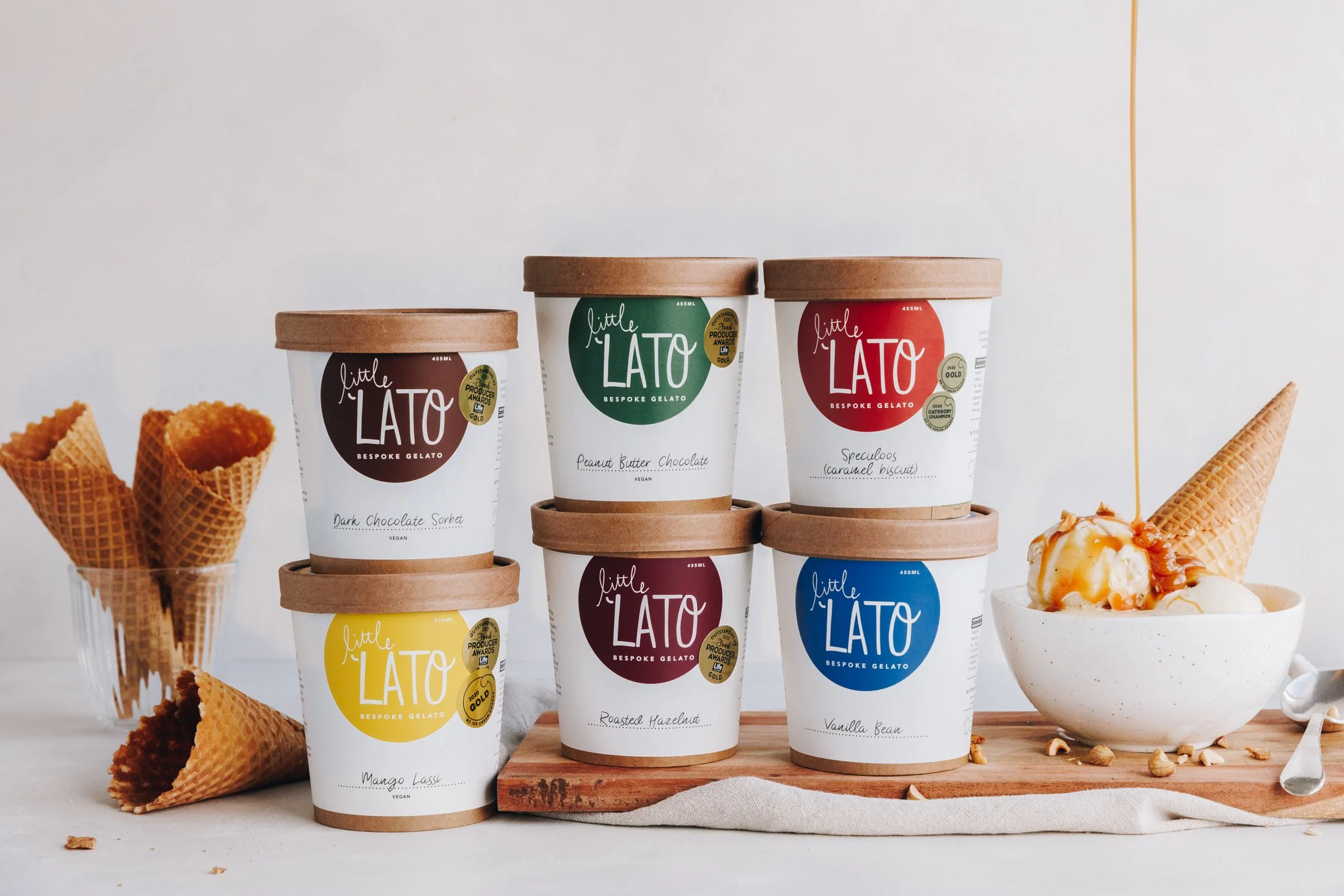 Collection of six pints of Little Lato gelato flavors arranged on a wooden stand, with waffle cones on the sides. Flavors include dark chocolate sorbet, peanut butter chocolate, speculoos caramel biscuit, roasted hazelnut, vanilla bean, and mango lassi. There is a bowl of ice cream with caramel sauce and wafer cones on the right, with some scattered nuts and crumbs.