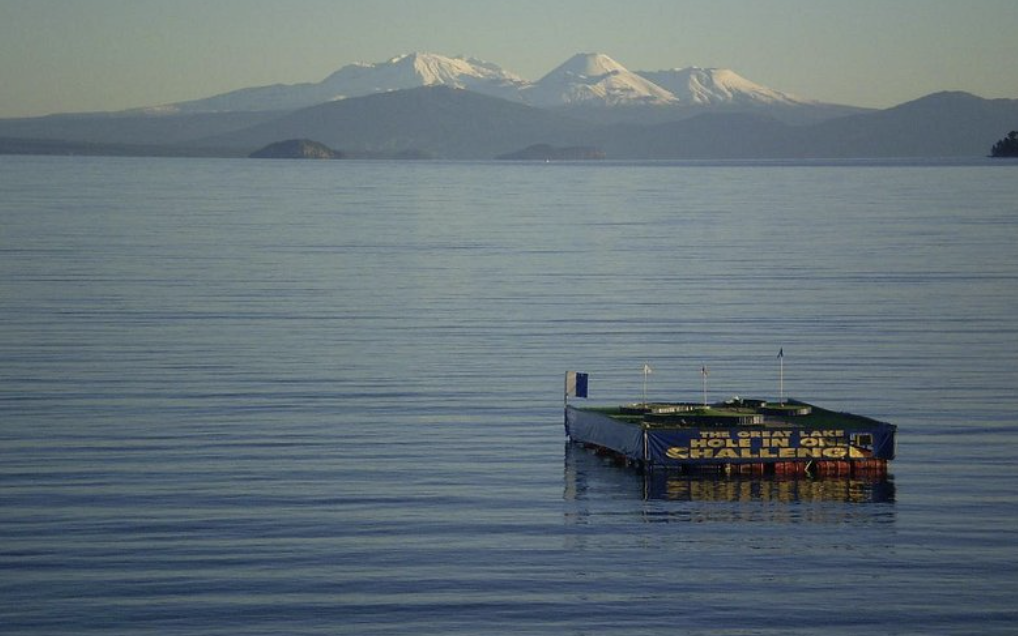 A floating golf challenge structure on a calm lake with snow-capped mountains in the background.