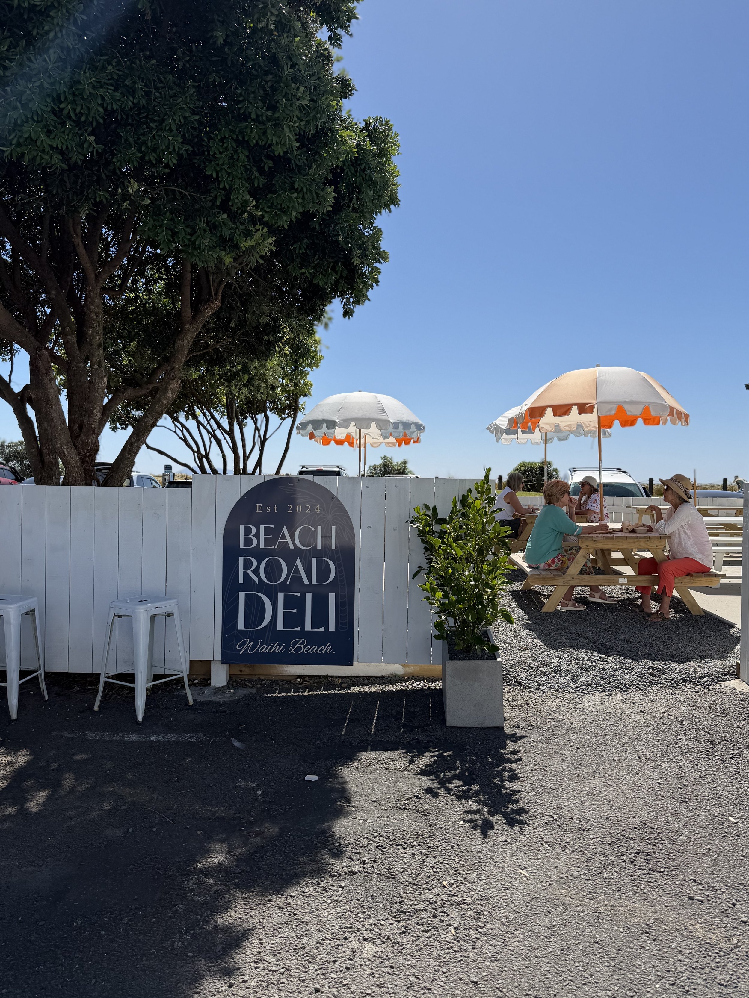 Outdoor seating area at a beachside restaurant or cafe with white fences, umbrellas, and people sitting at wooden tables, some wearing hats, under a large tree on a sunny day.