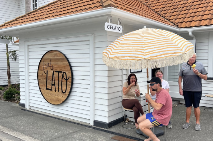 Group of four people outside an ice cream shop with a 'Gelato' sign hanging from the roof, sitting under a large striped umbrella, enjoying ice cream on a sunny day.
